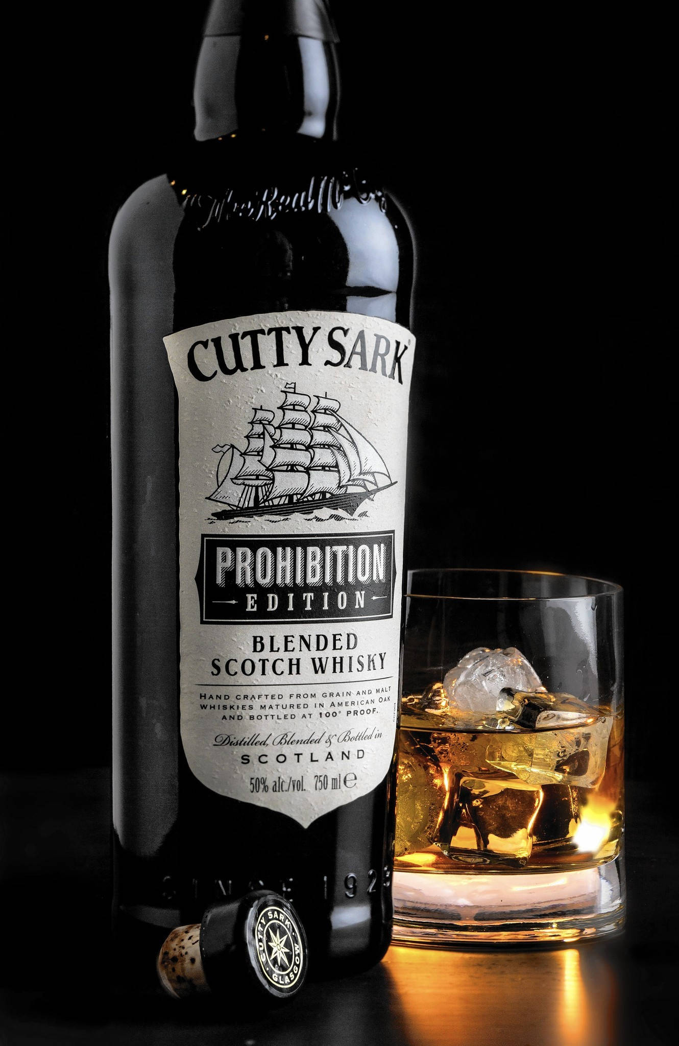 Cutty Sark 禁酒令版威士忌瓶 壁紙 — 一個纖細的黑色Cutty Sark Prohibition Edition威士忌酒瓶，旁邊放著一個裝滿琥珀色液體和冰塊的岩石杯，背景為深色。
