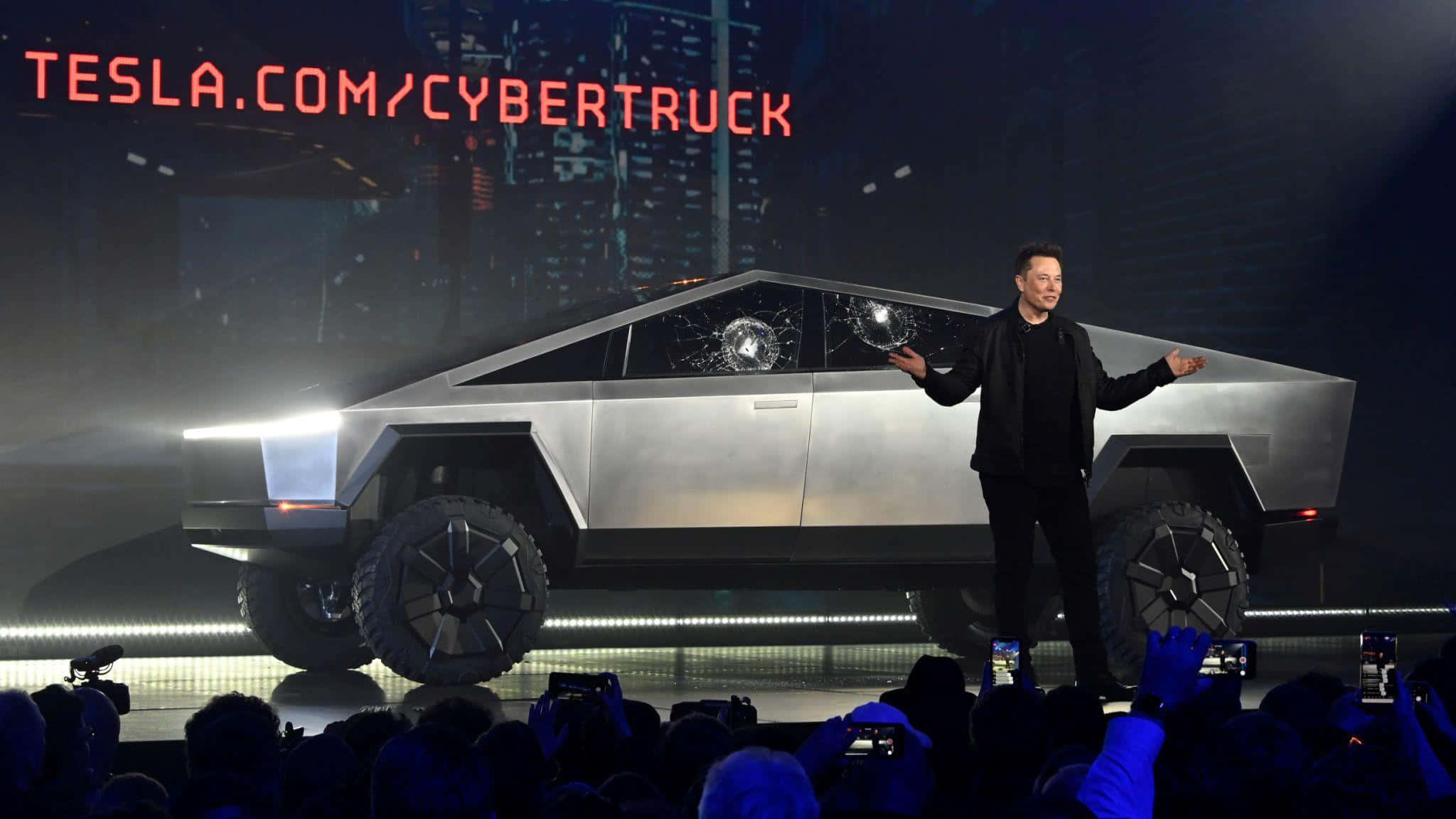 下一代交通已到來：特斯拉Cybertruck 壁紙 — 伊隆·馬斯克站在舞台上，在明亮的舞台燈光下，在一輛車窗破碎的銀色 Tesla Cybertruck 旁邊做手勢。