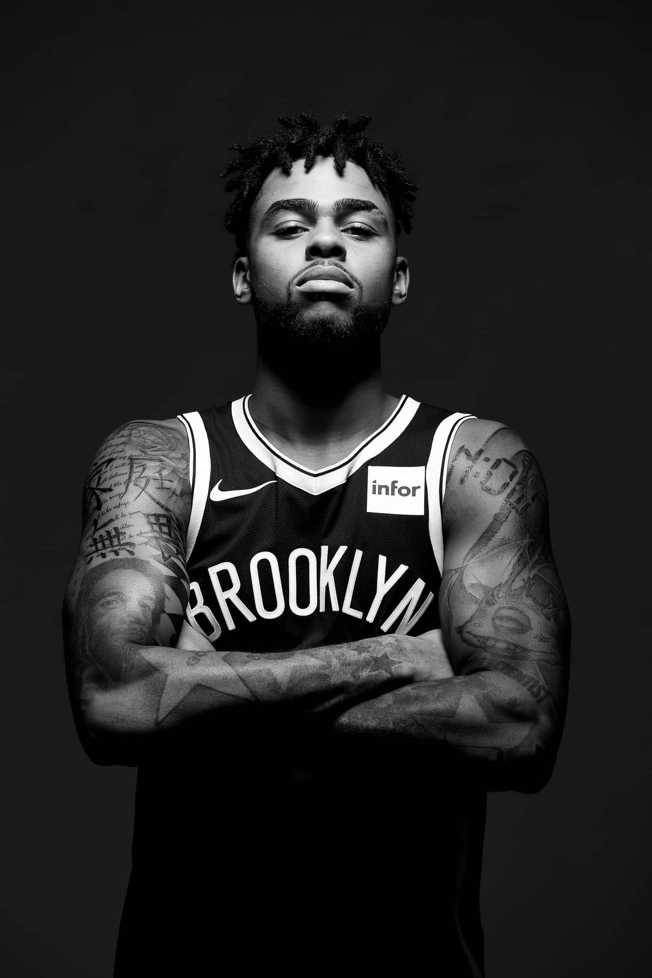 D'Angelo Russell低頭凝視 壁紙 — 一幅黑白肖像，展示一位紋身籃球運動員身穿布魯克林球衣，雙臂交叉站在深色背景前。