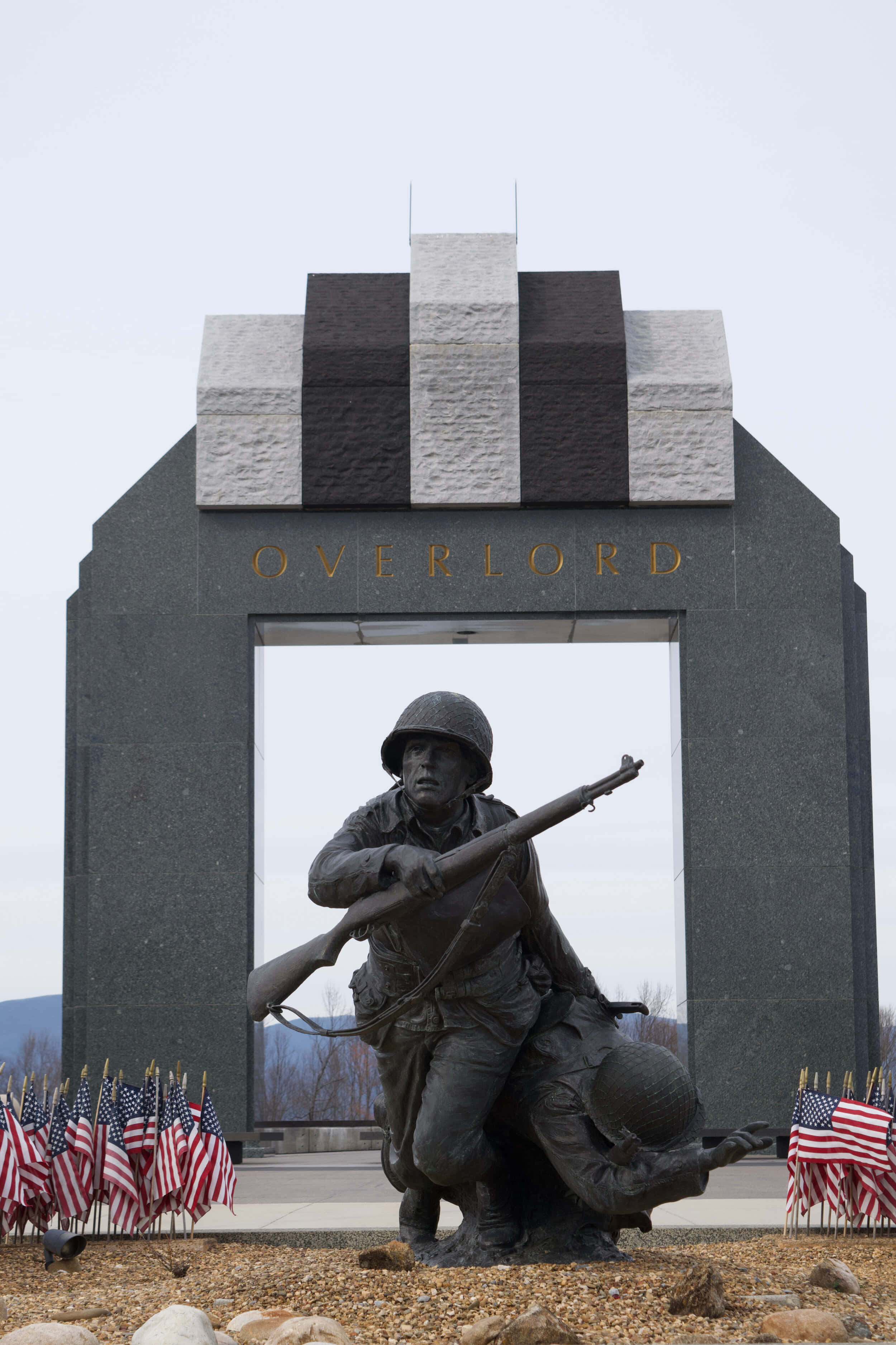 D-Day 登陸日，盟軍部隊駛向諾曼第灘頭 壁紙 — 一座莊嚴的兩名士兵作戰姿勢的青銅雕像矗立在刻有OVERLORD字樣的大型石拱門前，兩旁排列著美國國旗。