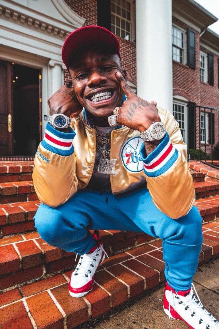 DaBaby 手機展示豪華腕錶 壁紙 — 一名微笑的男子身穿金色外套和藍色褲子，蹲在豪宅前的磚階上，戴著多隻手錶和紅色帽子。
