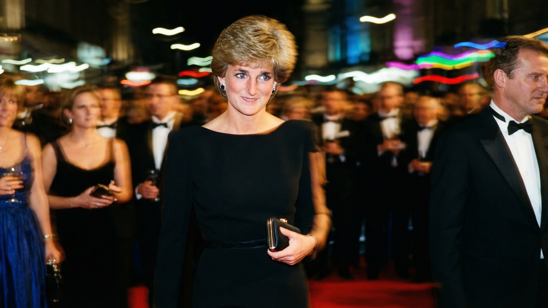 Princess Diana 桌布 壁紙 — 戴安娜王妃自信地走在紅地毯上，身穿黑色連衣裙，身後有模糊的賓客和五彩繽紛的燈光，參加一場華麗的晚間活動。