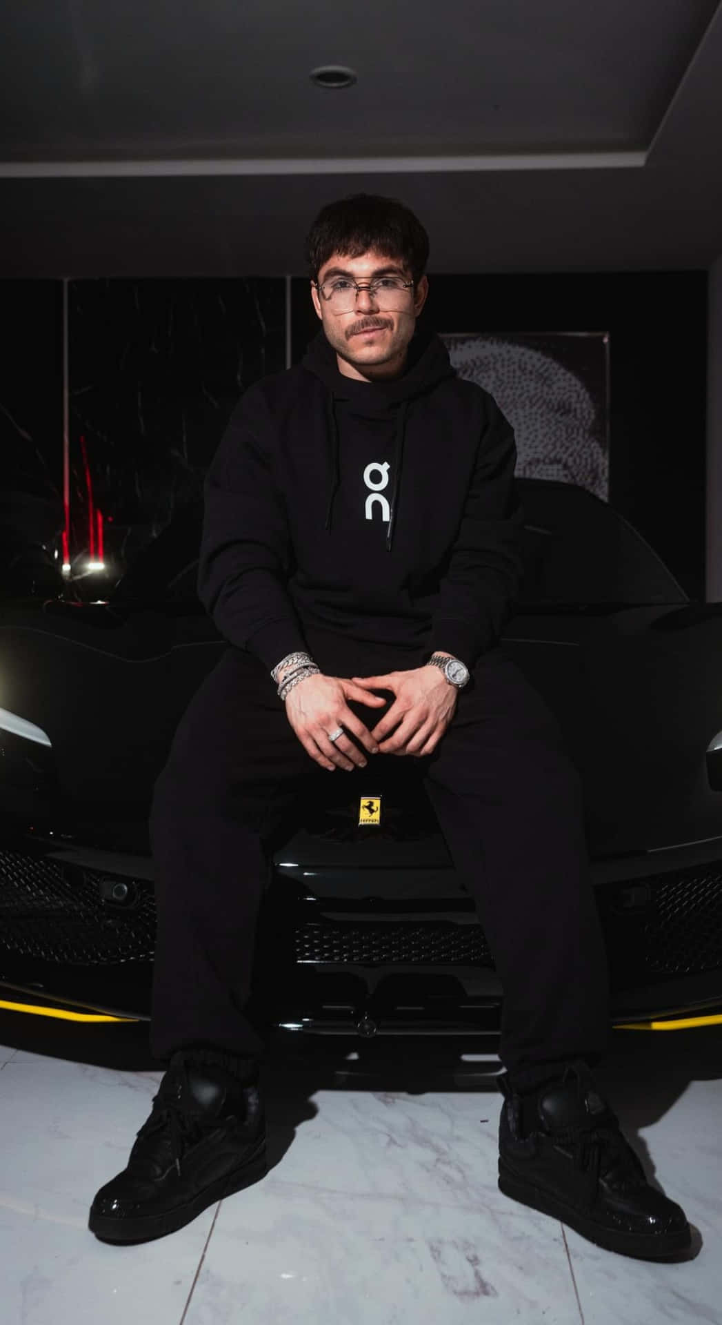  壁紙 — 一位戴眼鏡的年輕男子自信地坐在一輛時尚黑色跑車的引擎蓋上，身處燈光昏暗的現代車庫。