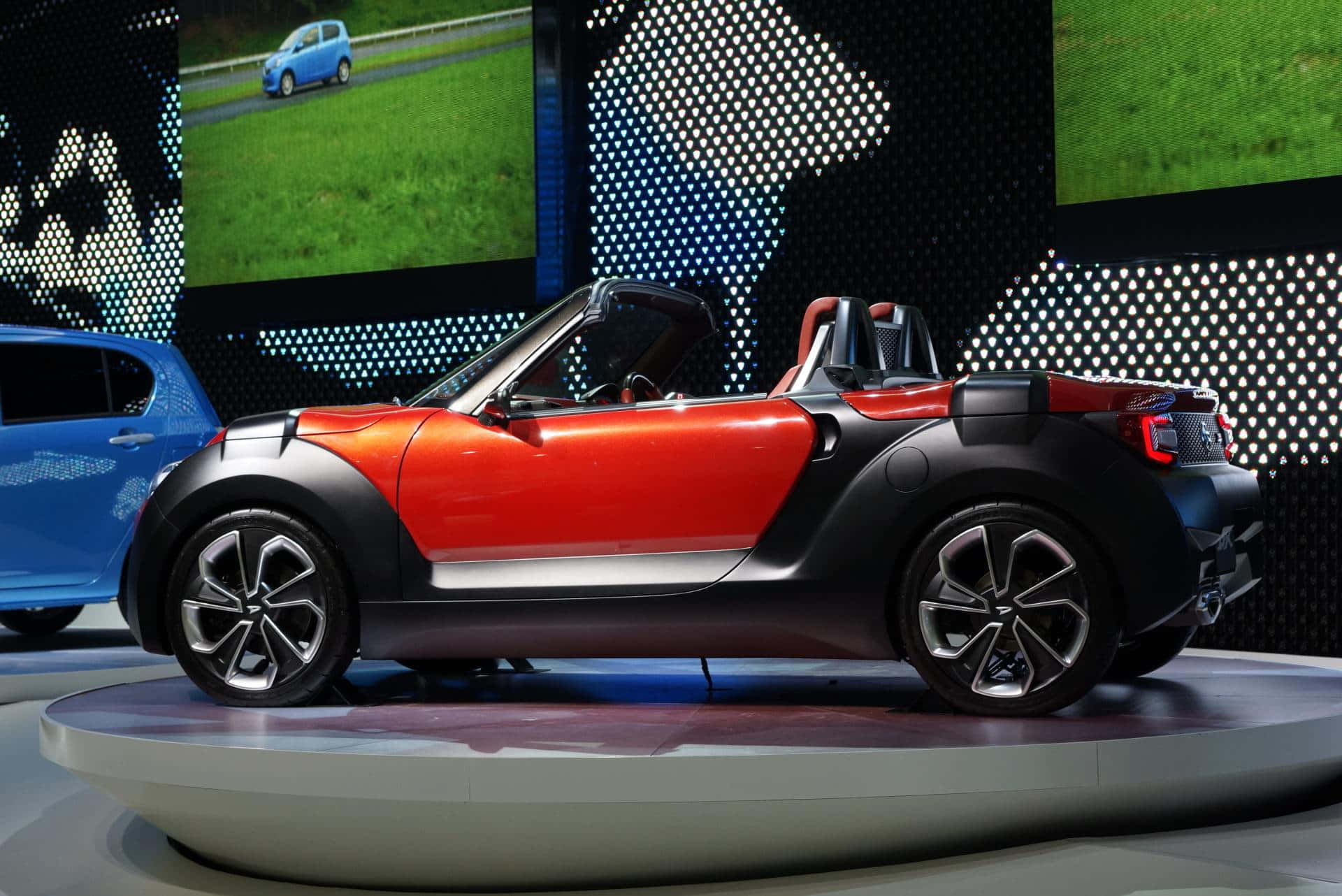 型格紅色Daihatsu喺開闊路上 壁紙 — 一輛鮮豔的紅色和黑色敞篷跑車在車展的圓形平台上展示，背景有數位螢幕。