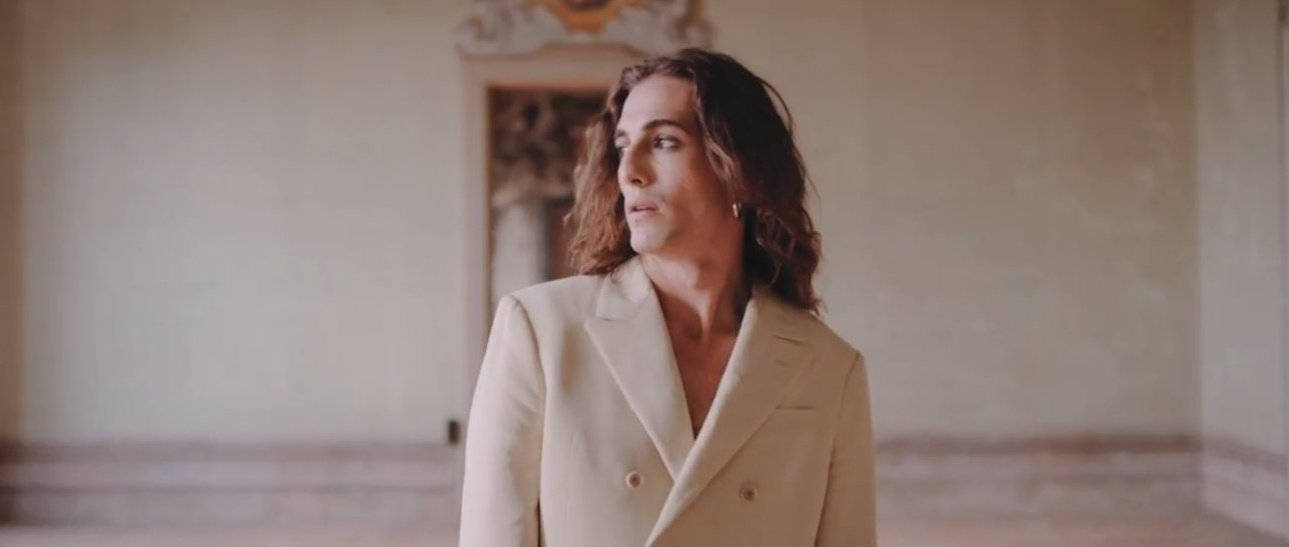 Damiano David 的美學氛圍 壁紙 — 一位擁有波浪形頭髮、身穿奶油色西裝外套的人，站在一個宏偉、光線柔和且裝飾華麗的房間裡。