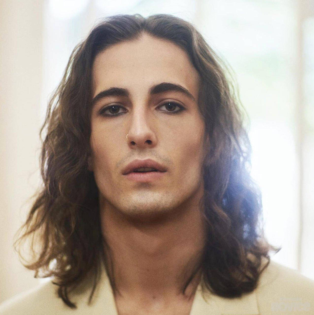 Damiano David 的天使氣質 壁紙 — 一位年輕男子的特寫肖像，他留著長長的波浪形棕色頭髮，戴著鼻環，在柔和自然光線下直視鏡頭。