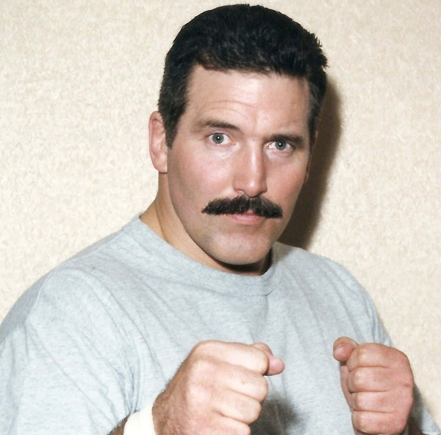 Dan Severn - 職業摔角的野獸 壁紙 — 一個留著濃密鬍鬚、眼神銳利的男人，握緊拳頭，對著有紋理的米色牆壁擺姿勢。