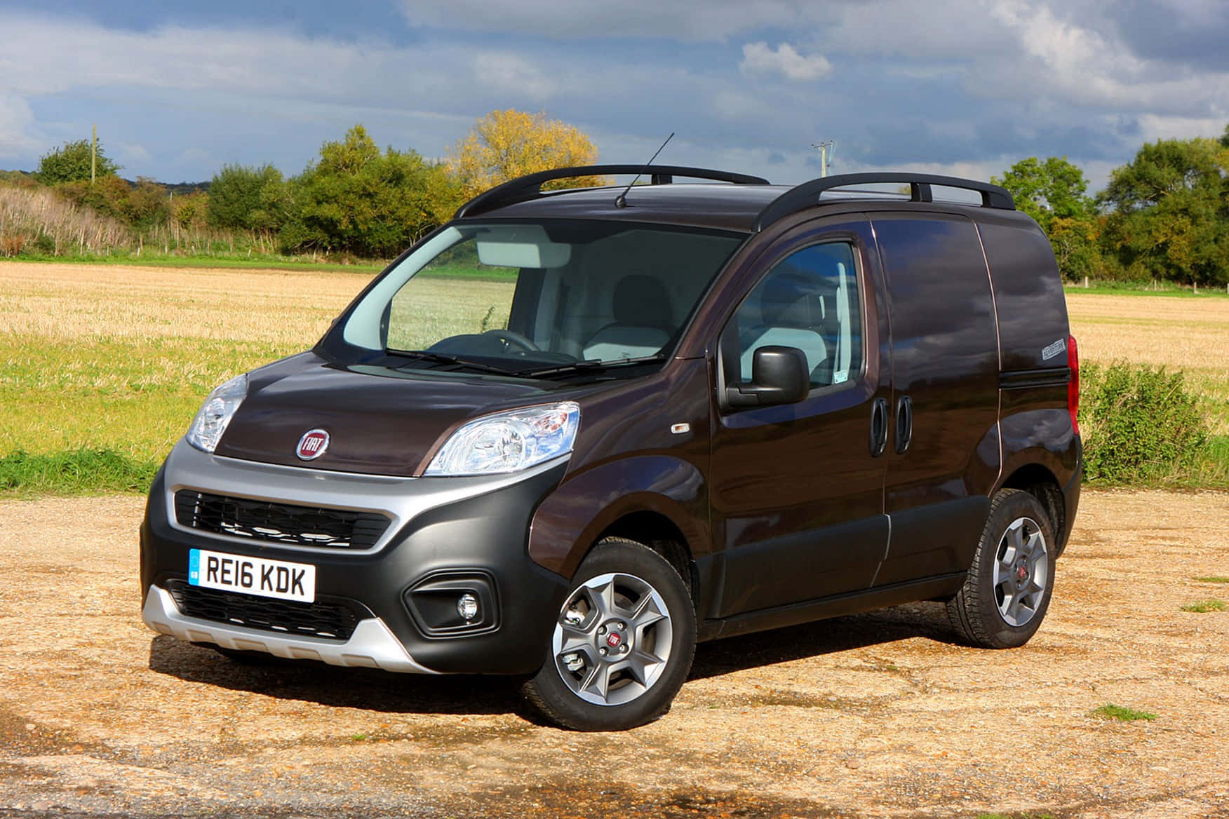 Fiat Fiorino 桌布 壁紙 — 一輛深啡色嘅Fiat客貨車停泊喺碎石場上，天空多雲，背景有綠色同黃色嘅樹木。