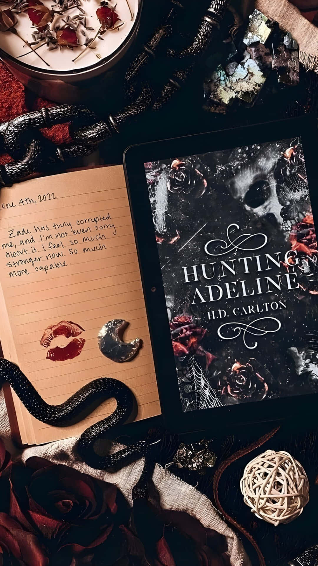  壁紙 — 一個黑暗哥德式靜物，展示了一部顯示「Hunting Adeline」的平板電腦，旁邊有一本帶有唇印的手寫日記，還有一條黑蛇盤繞在附近。