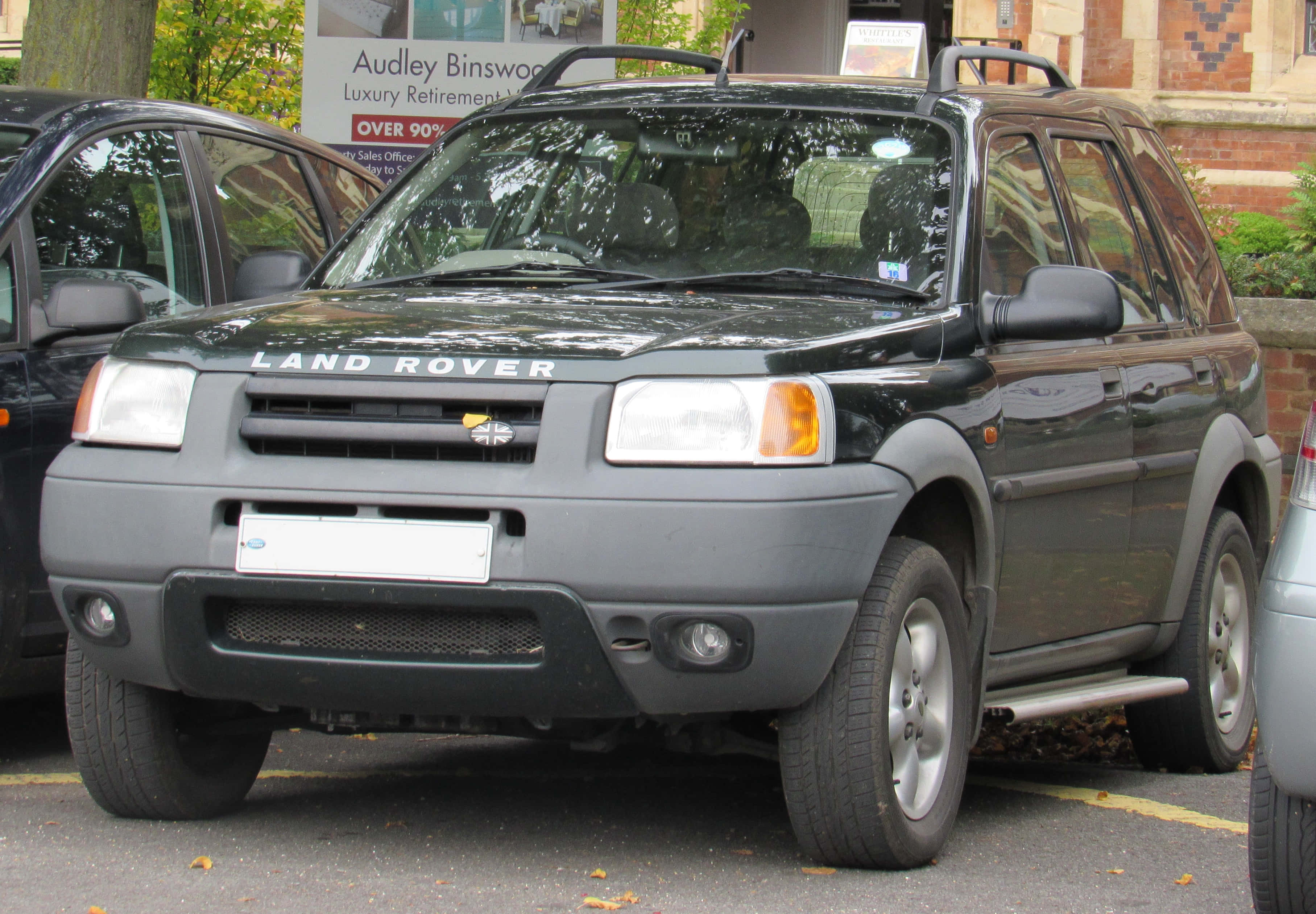 Land Rover Freelander 桌布 壁紙 — 一架深灰色嘅 Land Rover Freelander 泊喺一個落葉遍地嘅城市空地，旁邊有磚砌建築同其他汽車。