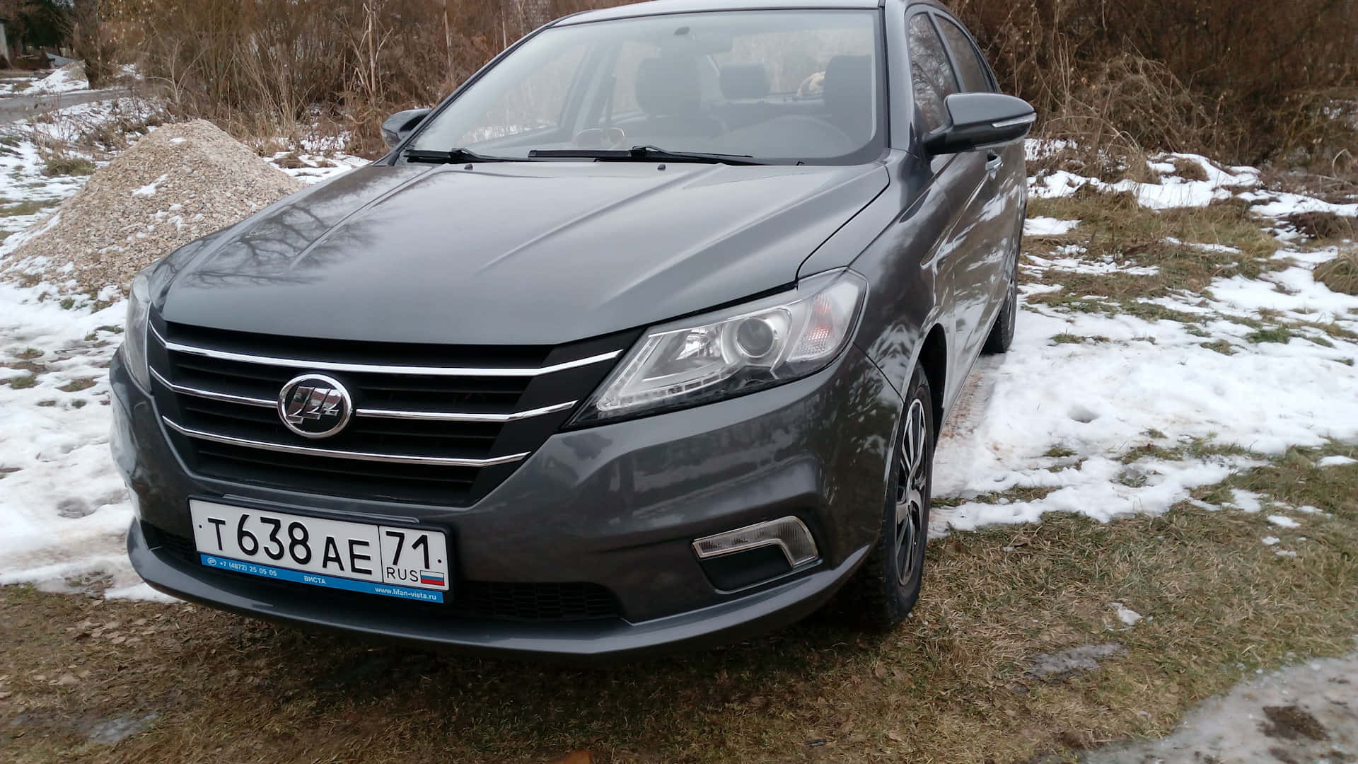  壁紙 — 一輛深灰色轎車停泊在積雪草地上，背景有光禿禿的灌木叢。
