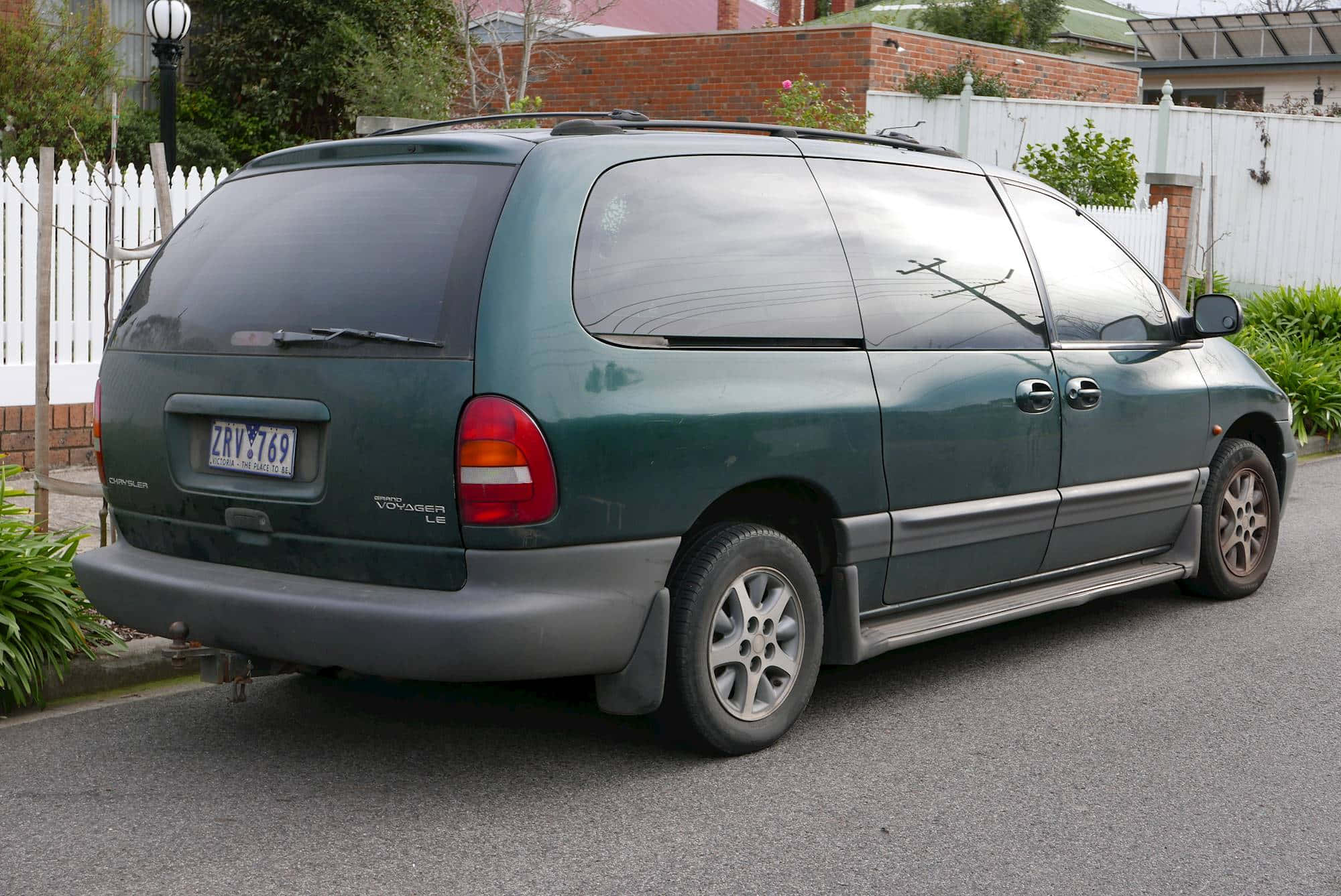 Chrysler Voyager 桌布 壁紙 — 一架深綠色嘅Chrysler Voyager小型貨車停泊喺住宅區街道旁邊，隔籬有白色籬笆同磚牆。