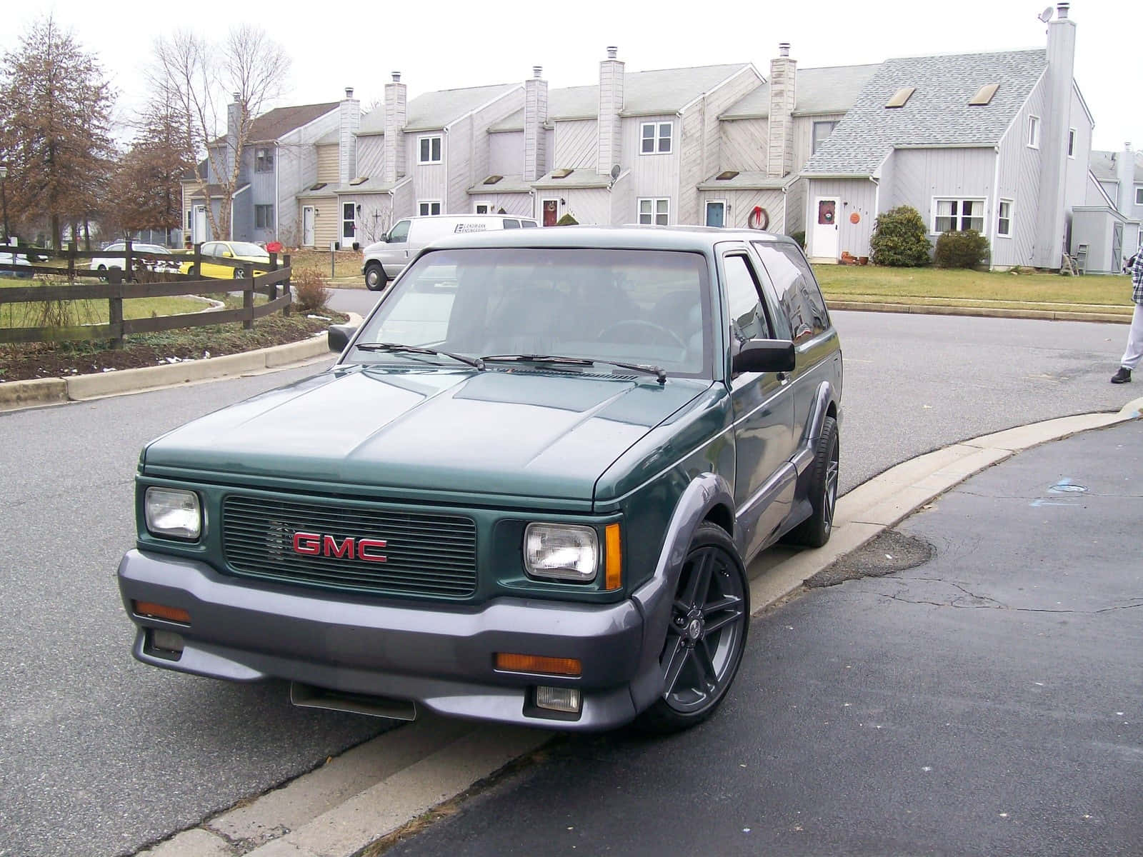 Gmc Typhoon 桌布 壁紙 — 一輛深綠色GMC Jimmy SUV停泊在住宅街道上，背景有郊區房屋和光禿禿的樹木。