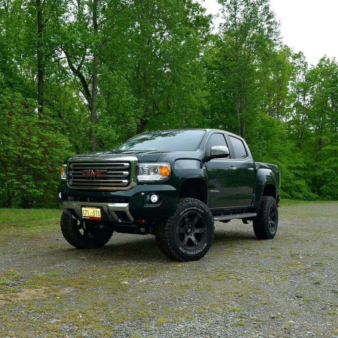 Gmc Canyon 桌布 壁紙 — 一輛深綠色GMC Sierra皮卡車，配備升高懸掛和黑色輪圈，停在碎石地上，周圍是茂密的綠樹。