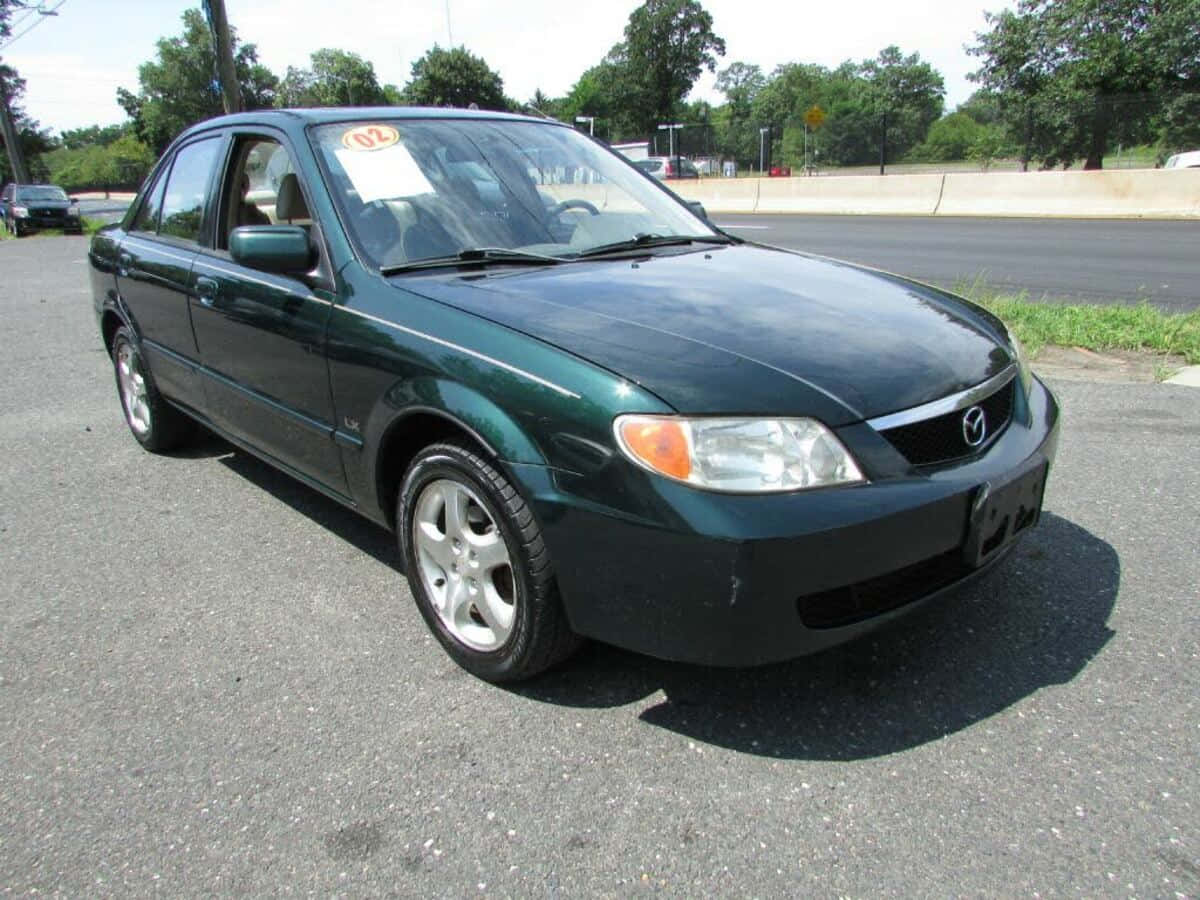 Mazda Protege 桌布 壁紙 — 一輛深綠色Mazda房車停在瀝青路上，背景有樹木和道路，在明亮的日光下。