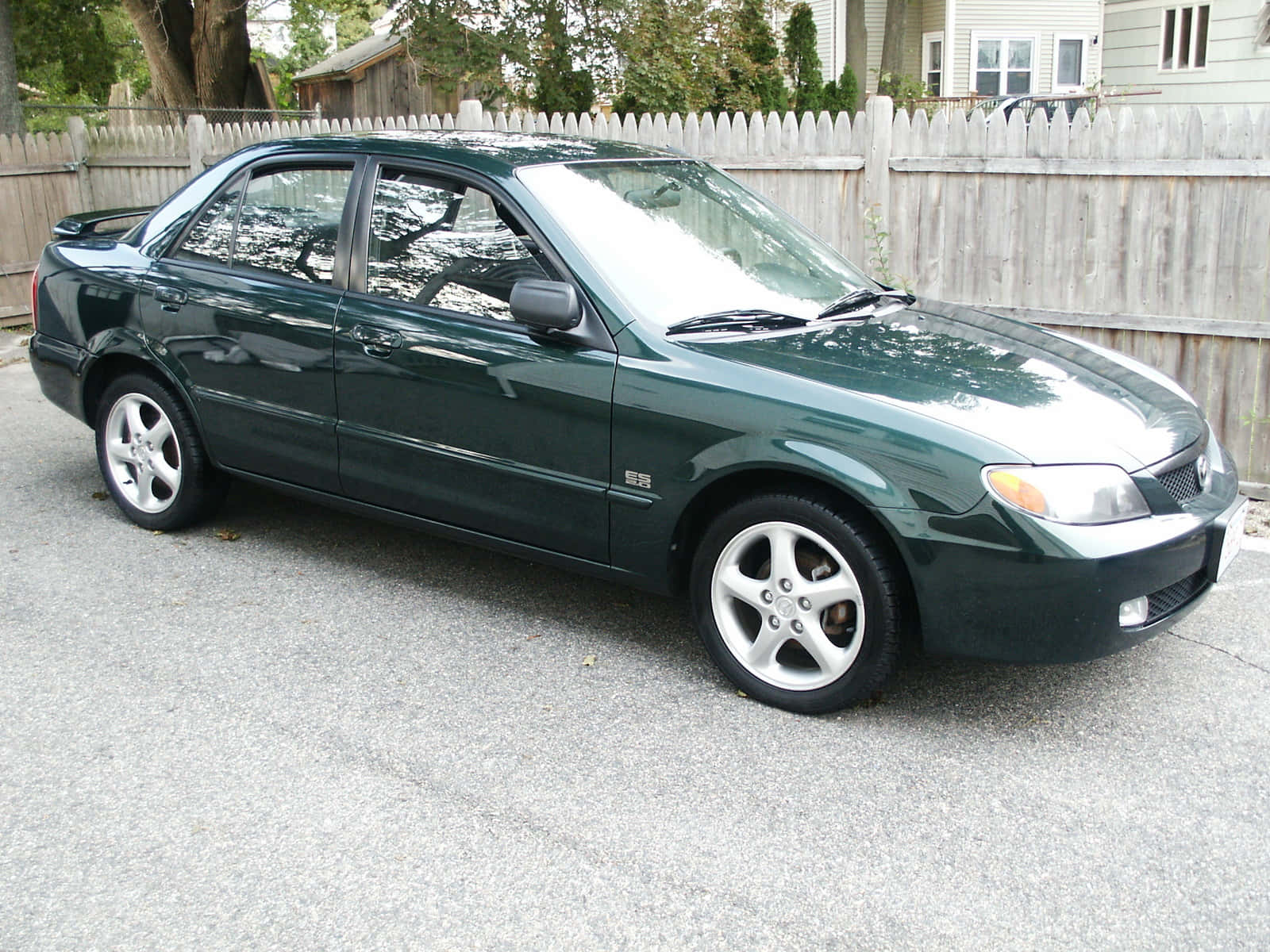 Mazda Protege 桌布 壁紙 — 一輛深綠色轎車配銀色輪圈，停泊在風化木柵欄前的碎石地上。