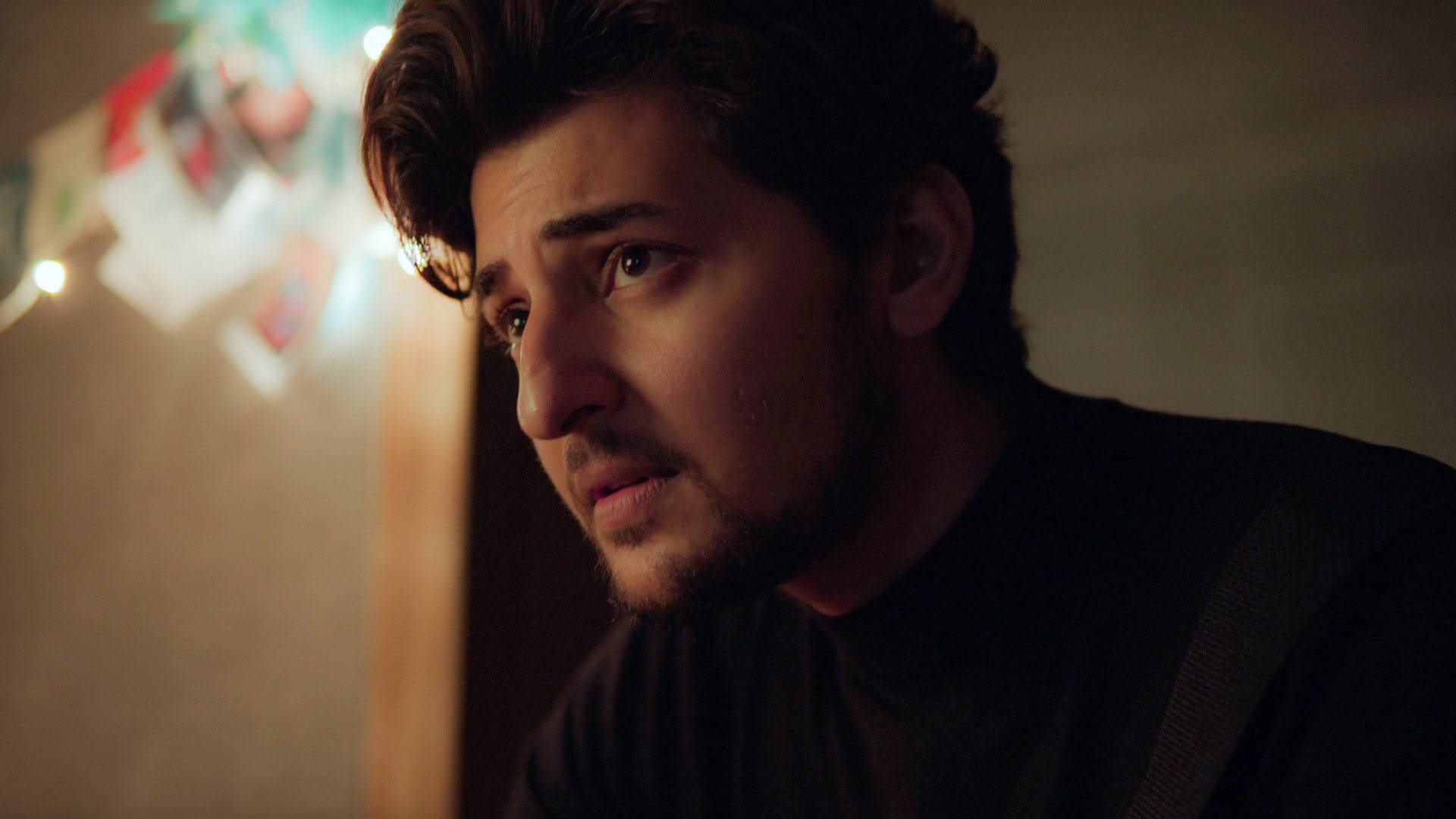 Darshan Raval 高畫質鄰家男孩 壁紙 — 一個深色頭髮和鬍鬚的男人在昏暗的房間裡沉思地凝視，背景有溫暖的串燈和模糊的照片。