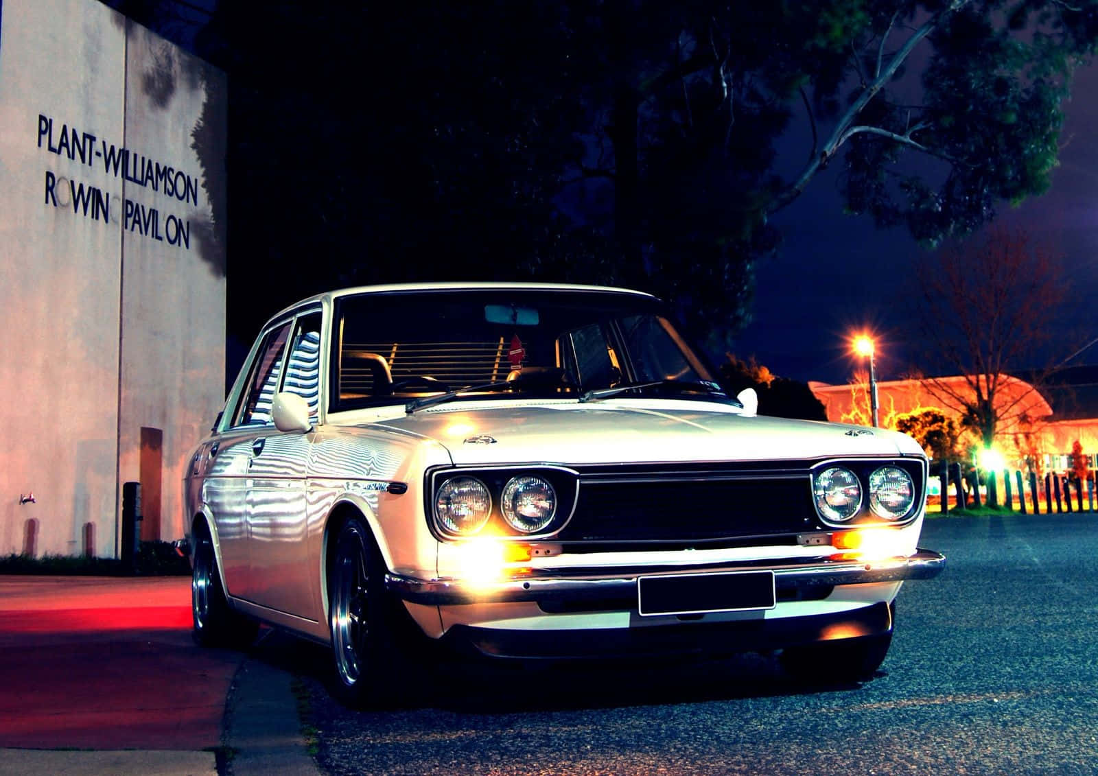 經典 Datsun 1600 在開放道路上駛過 壁紙 — 一輛銀色經典汽車在夜間停泊在設有照明標誌的建築物旁，在街燈下閃閃發光。