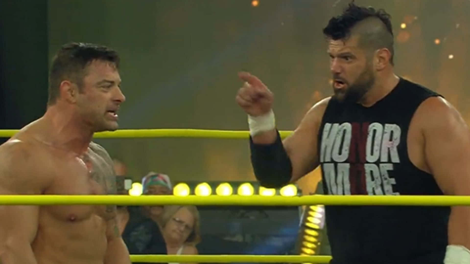 Davey Richards 與 Eddie Edwards 重聚 壁紙 — 兩名肌肉發達的摔角手在燈光明亮、帶有黃色繩索和閃爍燈光的擂台上互相怒視。