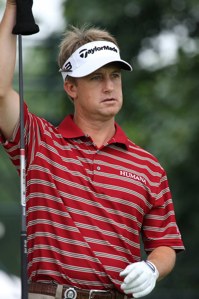 David Toms 全神投入高爾夫賽事 壁紙 — 一位專注的男性高爾夫球手，身穿紅色條紋襯衫和白色遮陽帽，在模糊的綠色背景下高舉球桿。