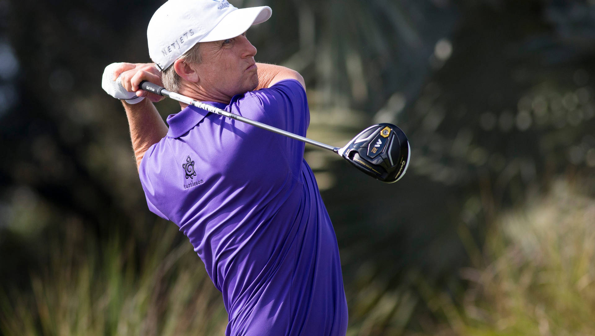 David Toms 高爾夫專注 壁紙 — 一位身穿紫色襯衫和白色帽子的高爾夫球手，在模糊的綠色背景下，專注地揮動一號木桿。