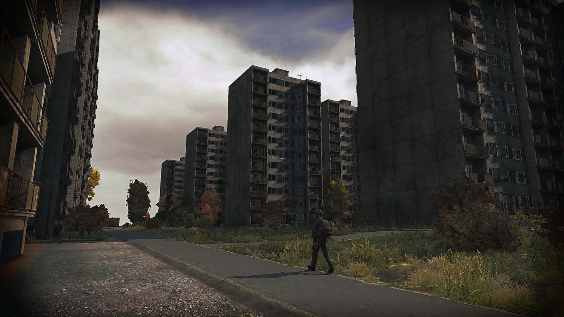 DayZ 1080p 背景：男子經過荒廢建築 壁紙 — 一個孤獨的身影在陰天之下，走在高聳風化的公寓大樓之間一條裂開的瀝青小徑上。