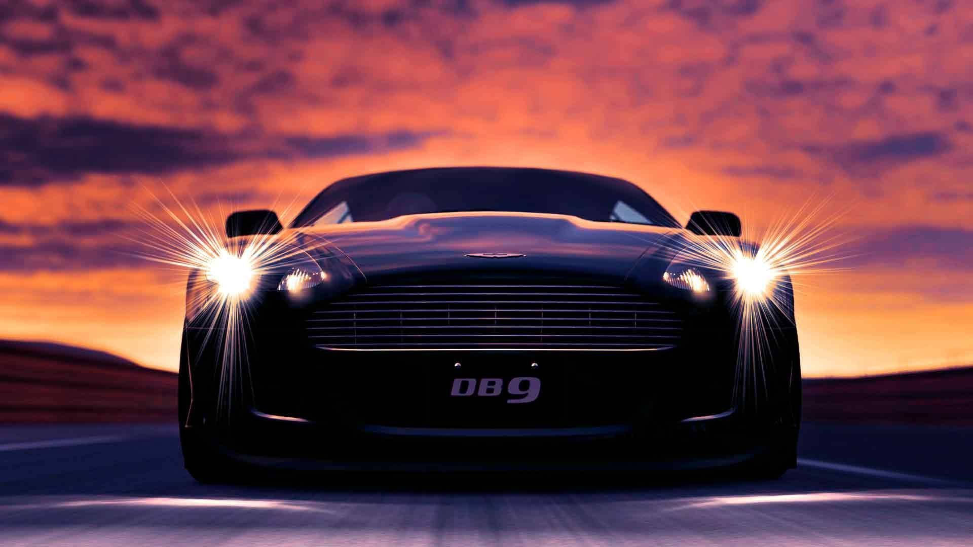 阿斯顿·马丁DB9 壁紙 — 一輛時尚的黑色 Aston Martin DB9 在熾熱的夕陽天空下，亮著車頭燈向觀眾疾馳而來。