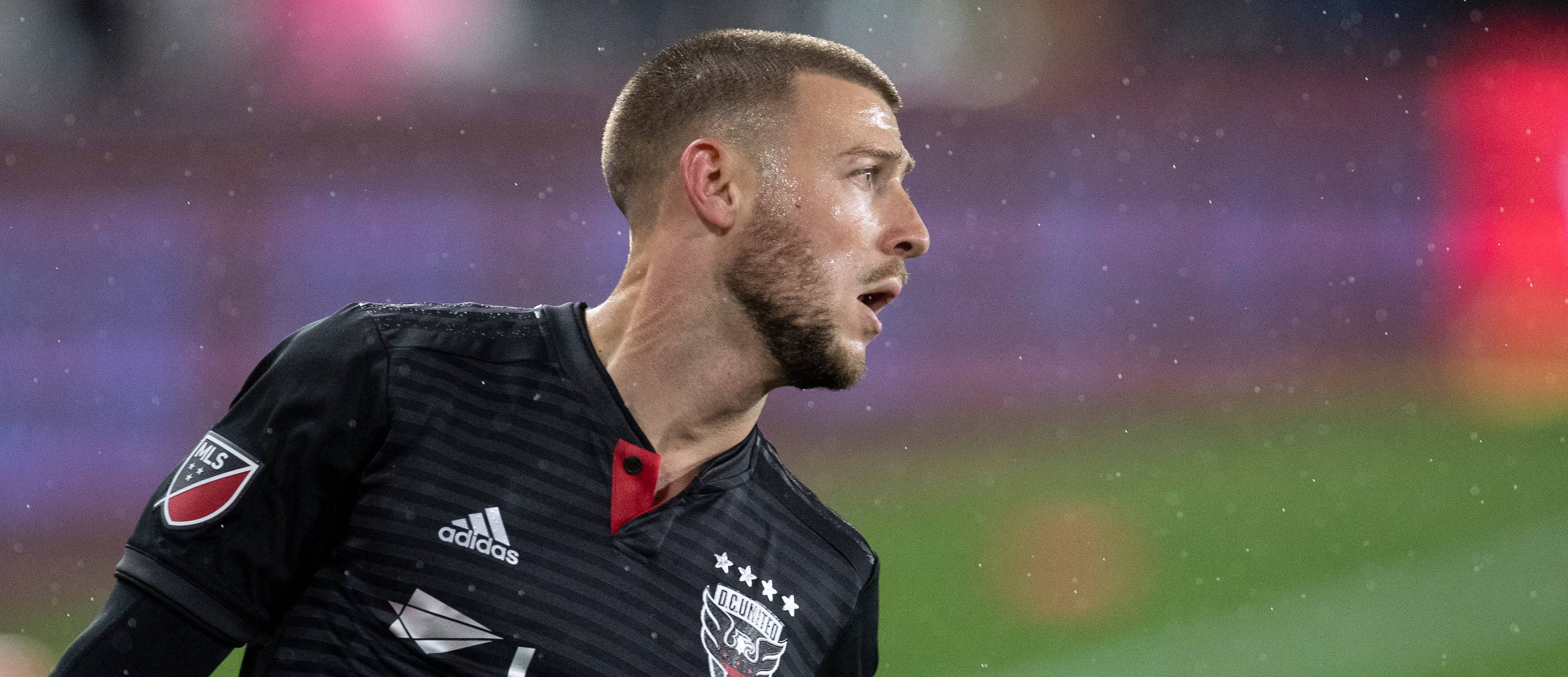 DC United Paul Arriola 在 MLS 賽事中堅持不懈 壁紙 — 一位身穿黑色球衣的專注足球員在雨夜比賽中，於球場燈光下沉重地呼吸。