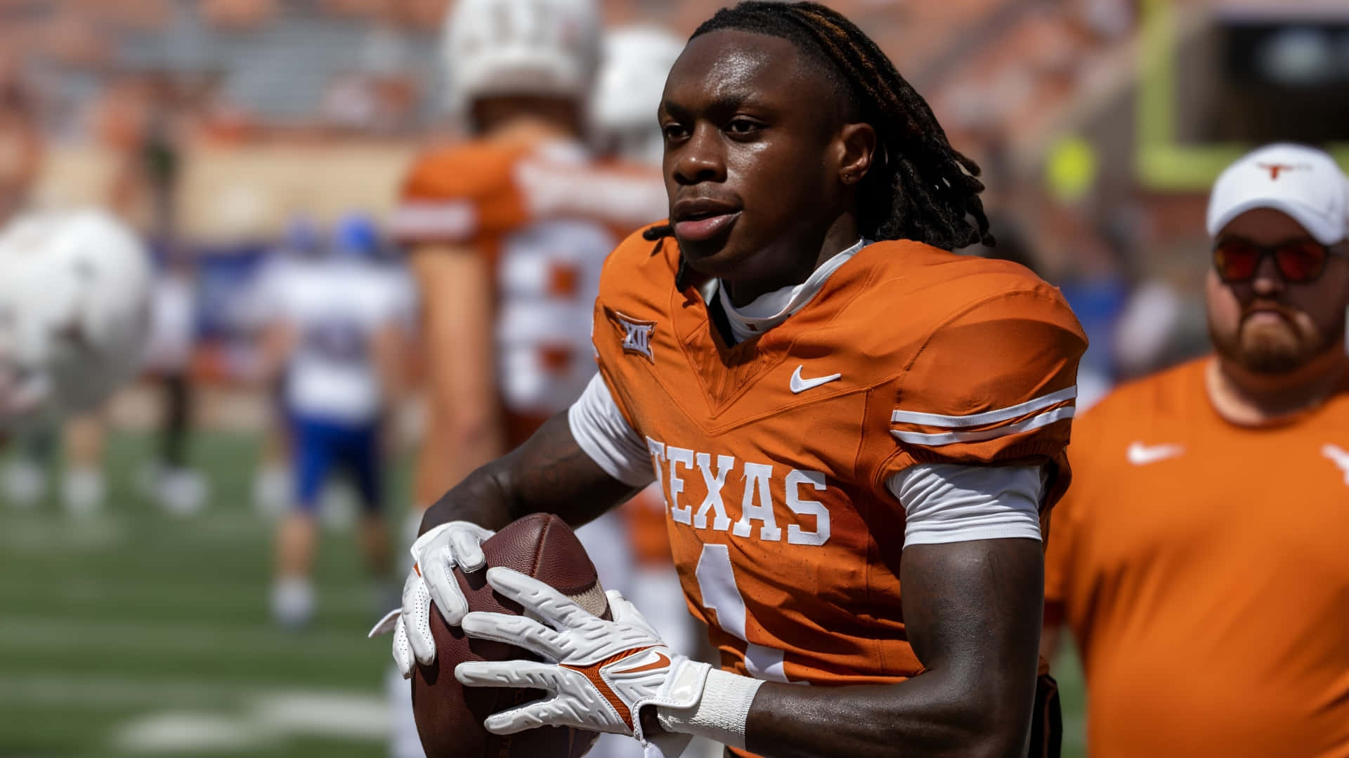  壁紙 — 一位身穿橙色球衣的德州美式足球員在陽光明媚的球場上抱著美式足球，背景有模糊的隊友。