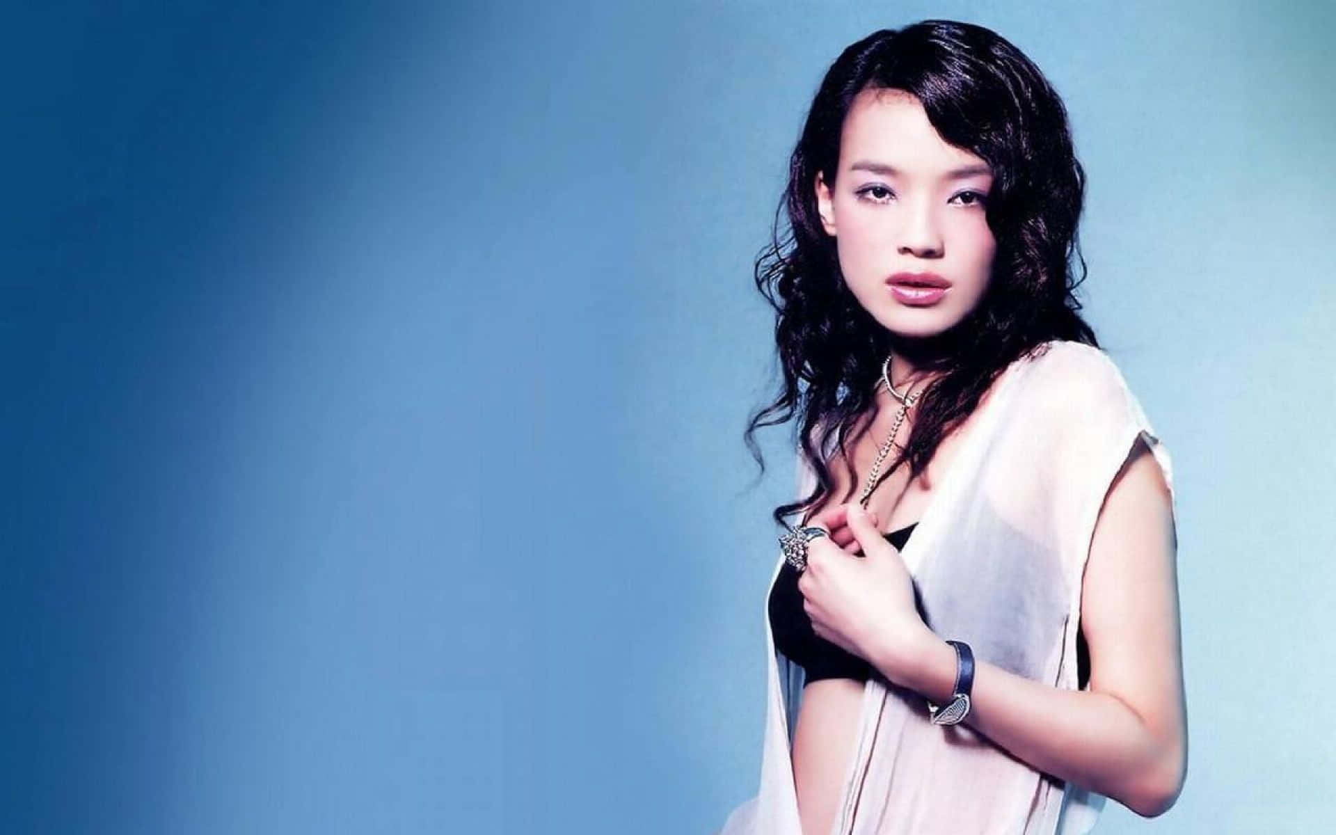 Shu Qi 桌布 壁紙 — 一位深色波浪髮的女性在柔和的藍色漸變背景前擺姿勢，身穿黑色上衣外搭透明白色上衣。