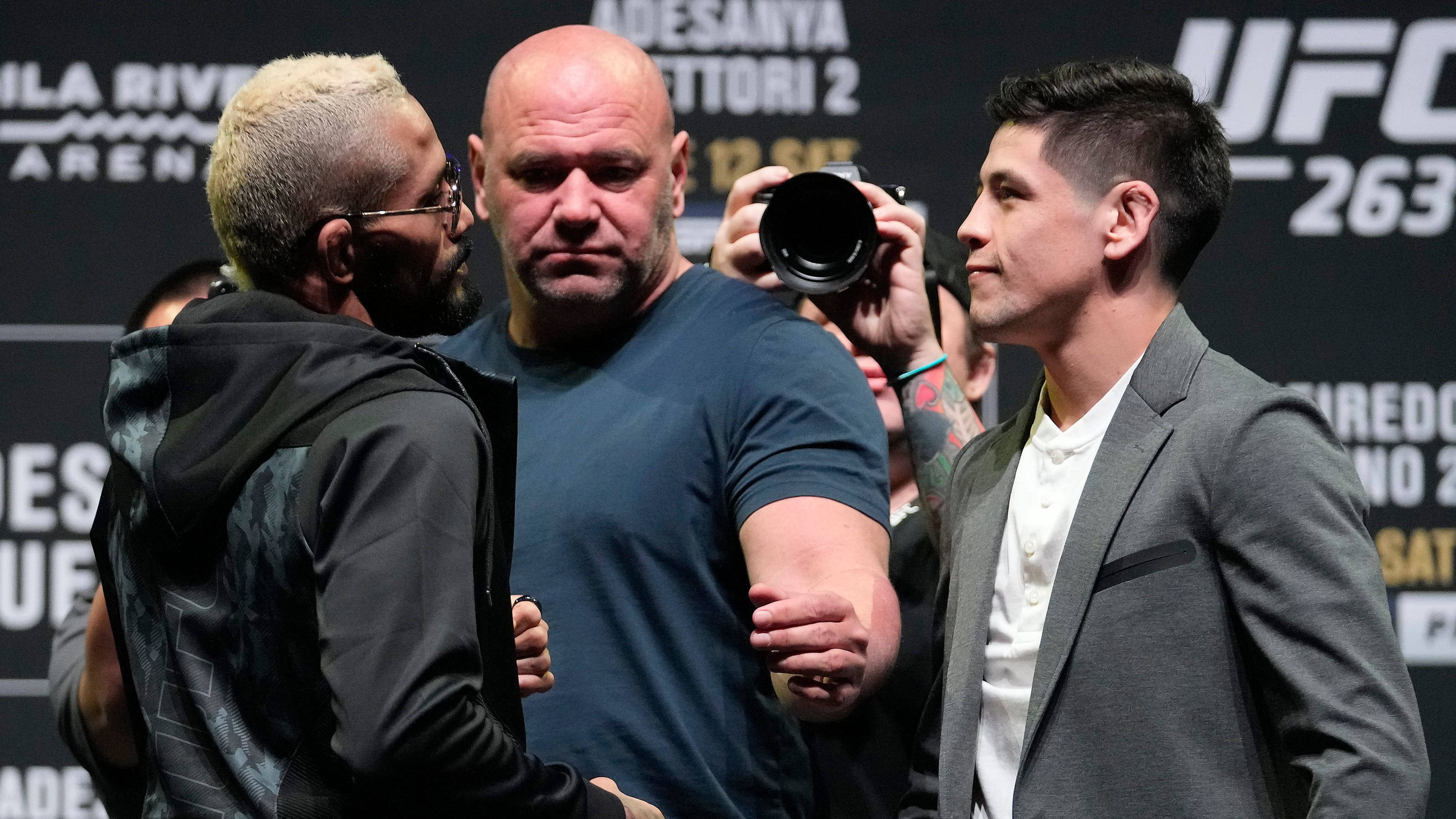 Deiveson Figueiredo 與 Brandon Moreno 面對面 壁紙 — 兩名UFC拳手在記者會上對峙，中間有裁判，攝影師捕捉緊張時刻。