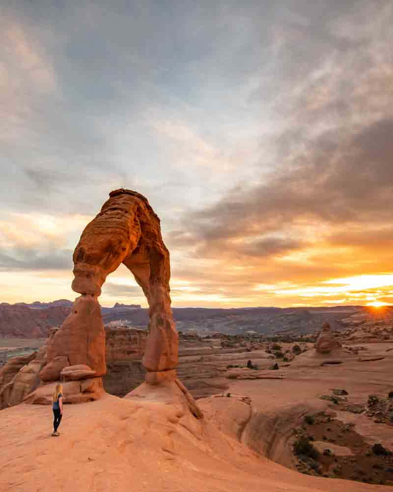 「日落時分的 Delicate Arch 壯麗景觀」 壁紙 — 一個孤獨的人站在雄偉的精緻拱門下，夕陽將溫暖的金色光芒灑在沙漠峽谷上。