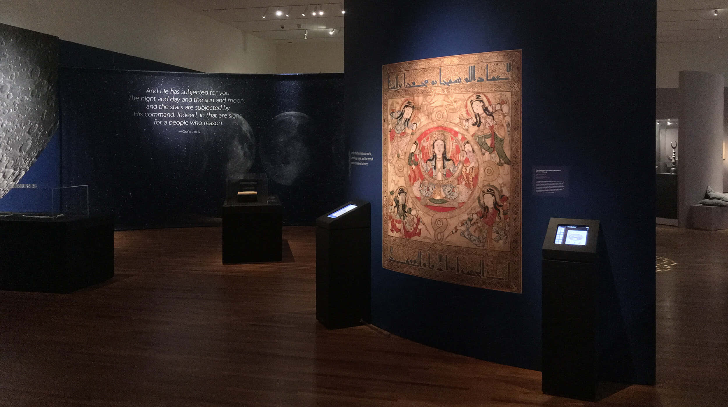 Aga Khan Museum 桌布 壁紙 — 一個燈光昏暗的博物館畫廊展示了一本大型華麗的伊斯蘭手稿，上面有天空圖案，背景是深藍色的牆壁，帶有月亮圖像和阿拉伯文字。
