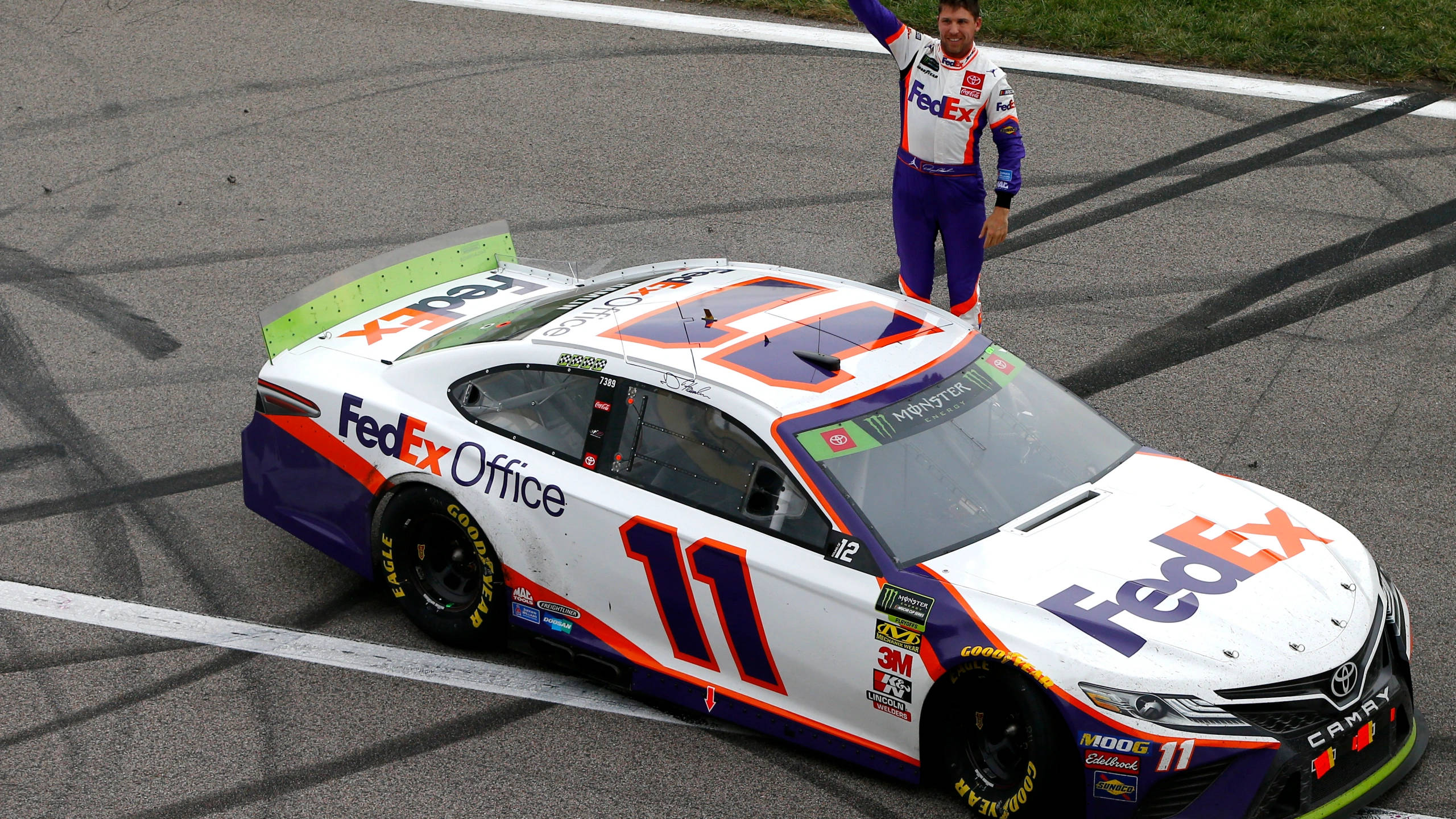 Denny Hamlin 與賽車合影 壁紙 — 一名身穿FedEx制服的NASCAR車手，在他白色、紫色和橙色的#11 FedEx Office賽車頂上慶祝，背景是賽道。