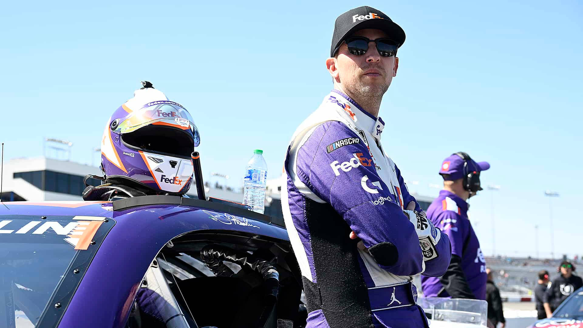 身穿紫色制服的NASCAR車手站在賽道上的賽車旁邊。