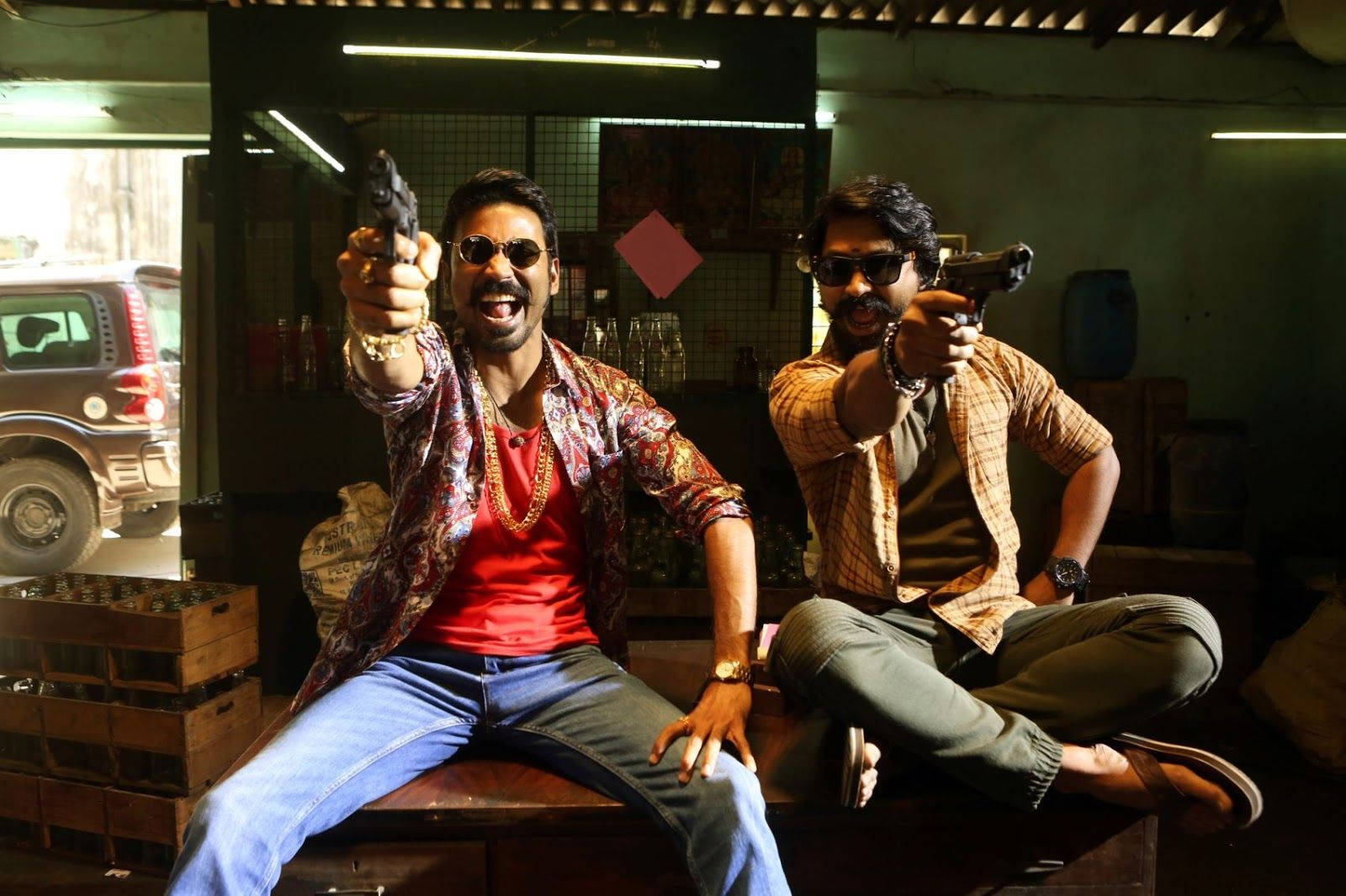 Dhanush 飾 Maari 2 指槍搞笑照 壁紙 — 兩個戴住太陽眼鏡嘅男人喺雜亂昏暗嘅車庫入面，帶住兇猛嘅笑容瞄準手槍。
