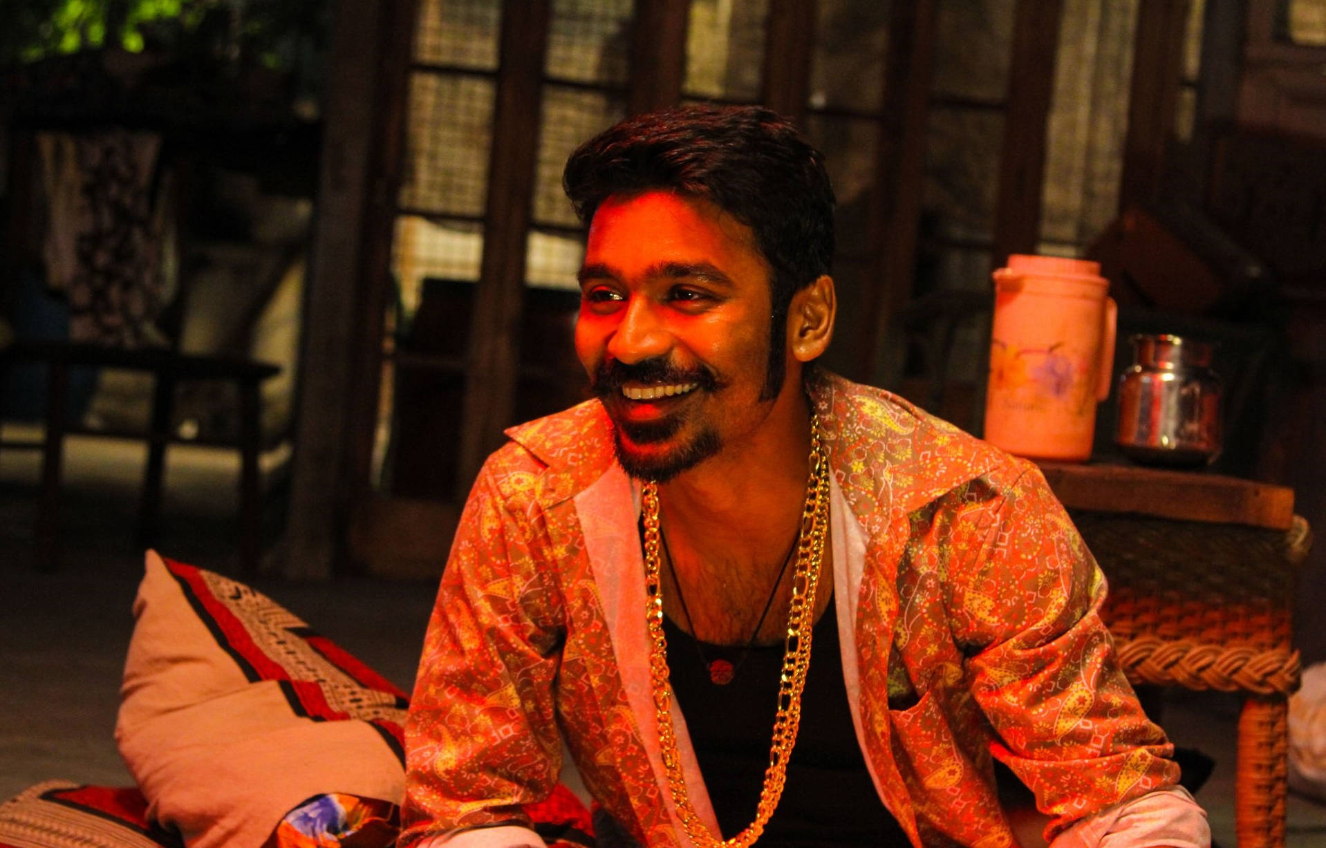 Dhanush 飾演 Maari 笑容滿面 壁紙 — 一名身穿鮮豔橙色圖案外套的男子，在溫暖的紅色燈光下，於鄉村風格的室內環境中溫暖地大笑。