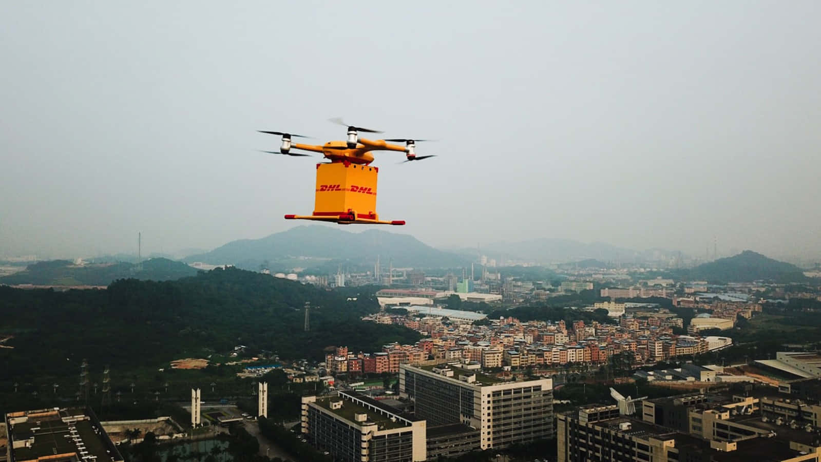 橙色DHL送貨無人機飛越城市，背景有山丘。