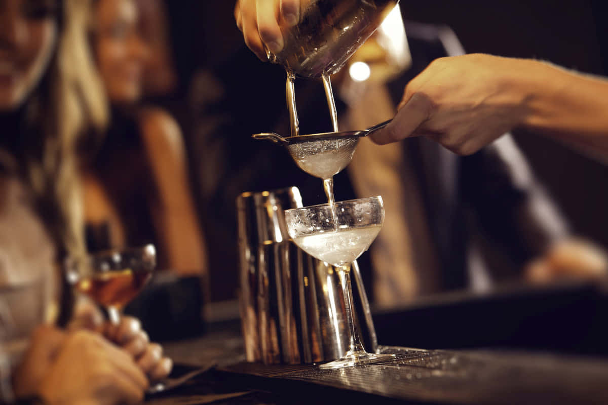  壁紙 — 調酒師將冰凍雞尾酒透過隔冰器倒入碟形杯，而顧客在昏暗的酒吧中手持飲品。