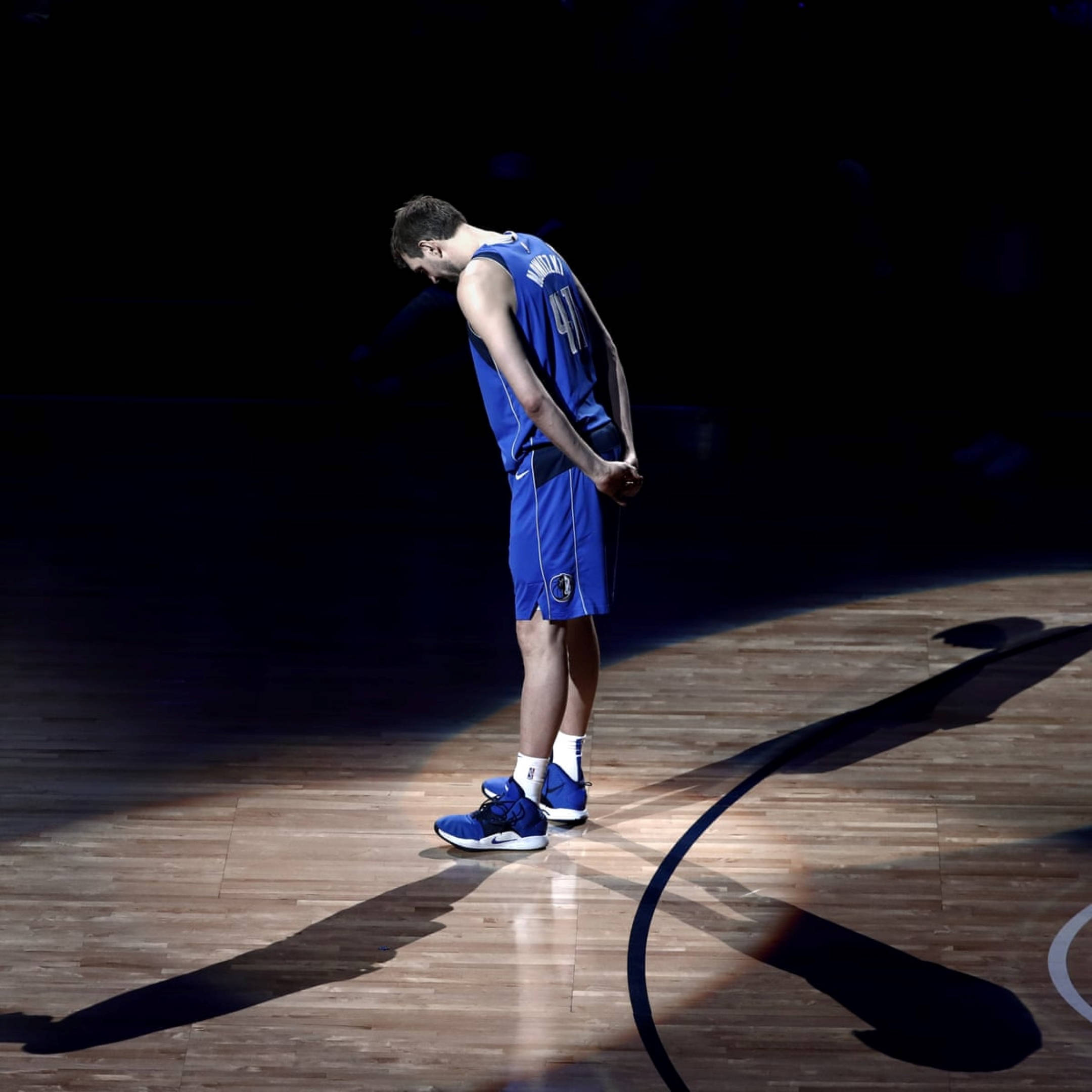 Dirk Nowitzki 2019退休賽事 壁紙 — 一名身穿藍色球衣的籃球運動員獨自站在球場上，在聚光燈下低頭沉思。
