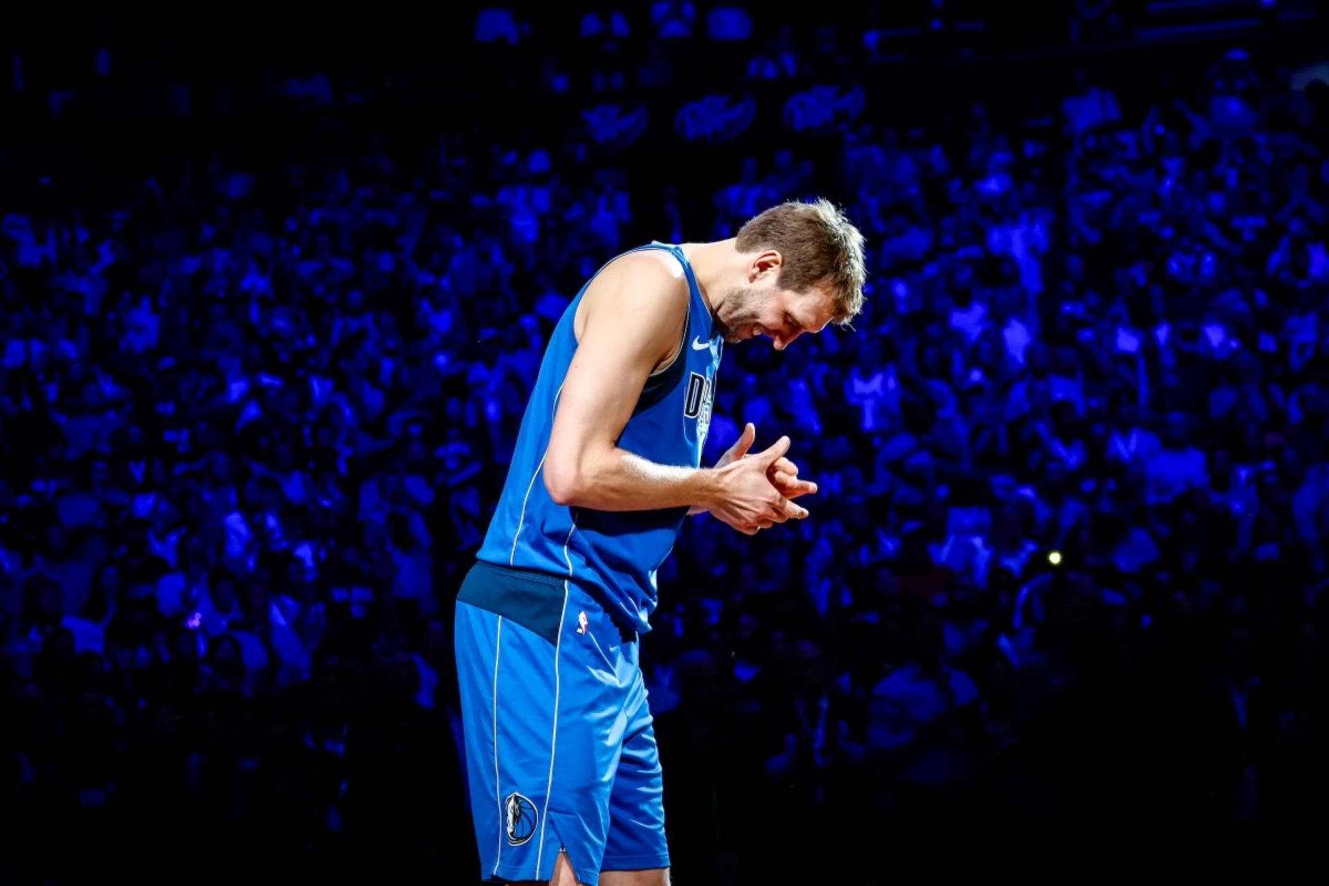 Dirk Nowitzki最後一場主場賽事 壁紙 — 一名身穿藍色球衣的籃球運動員在深藍色燈光下的人群中低頭合十。