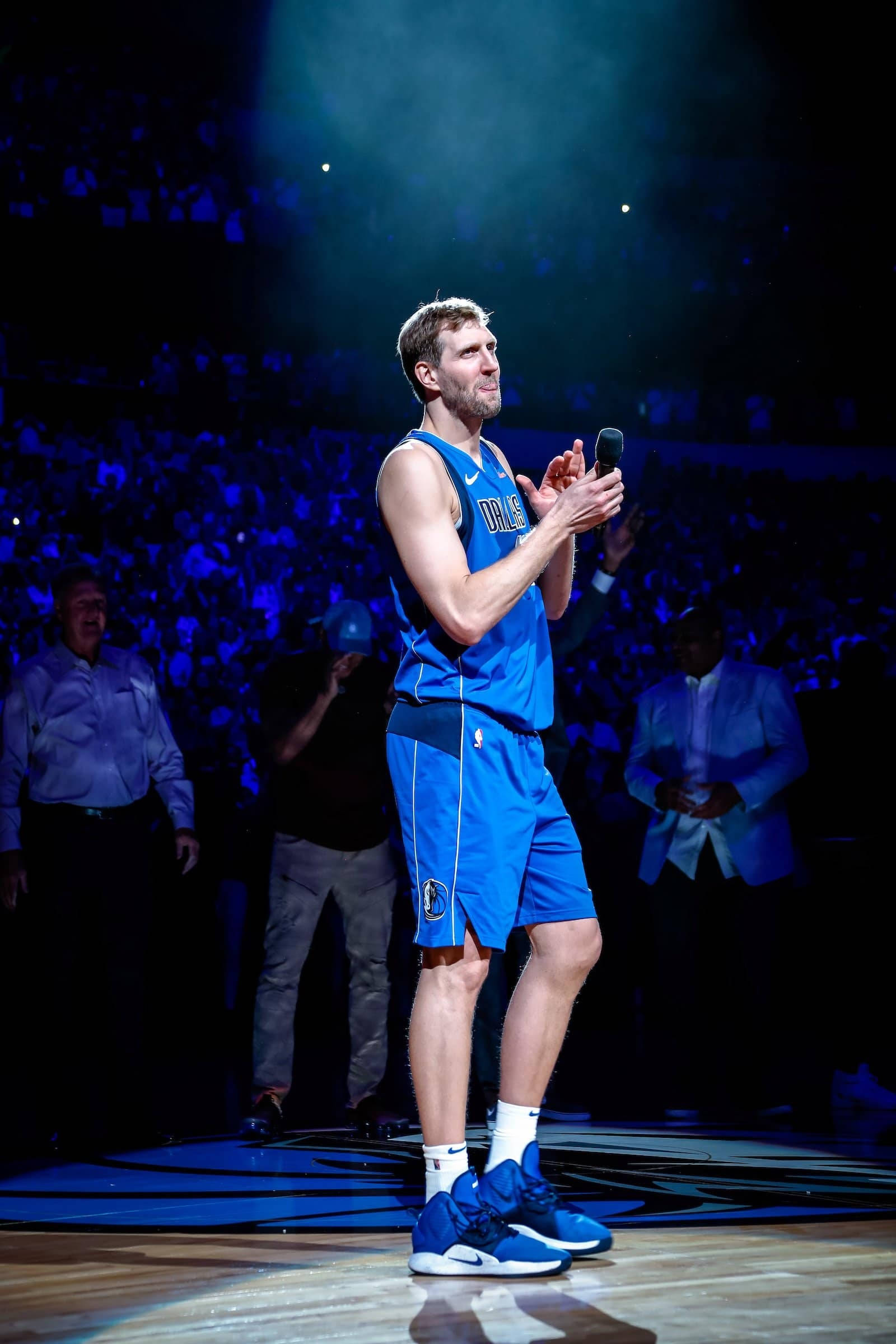 Dirk Nowitzki致辭時刻 壁紙 — 一名身穿藍色球衣的籃球運動員在明亮的舞台燈光下於球場上拍手，身後有模糊的人群。