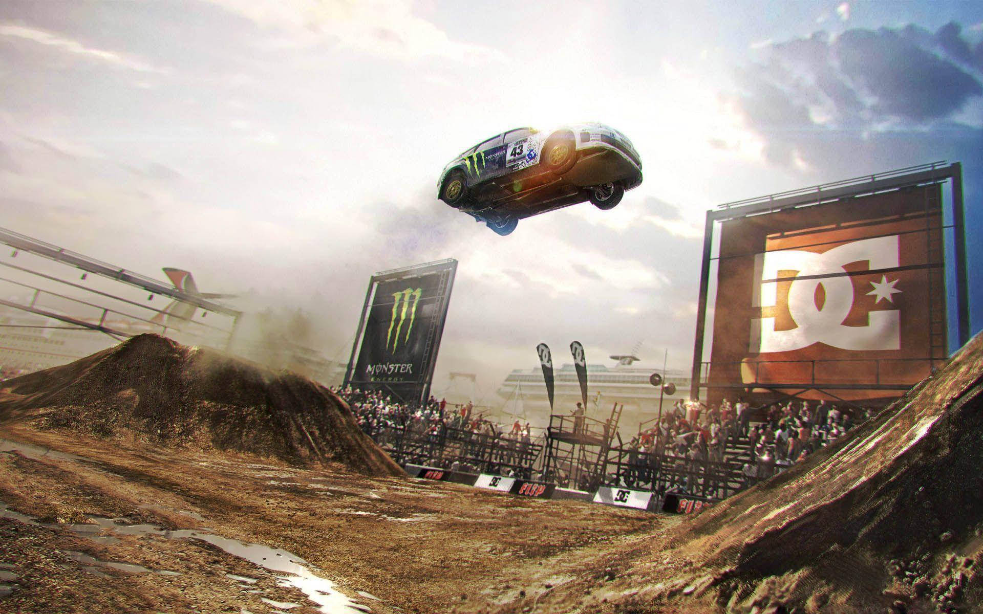 Dirt 3 車輛空中飛躍 壁紙 — 一輛拉力賽車在泥濘的土路上空飛馳，觀眾在陰天下的看台上觀看。