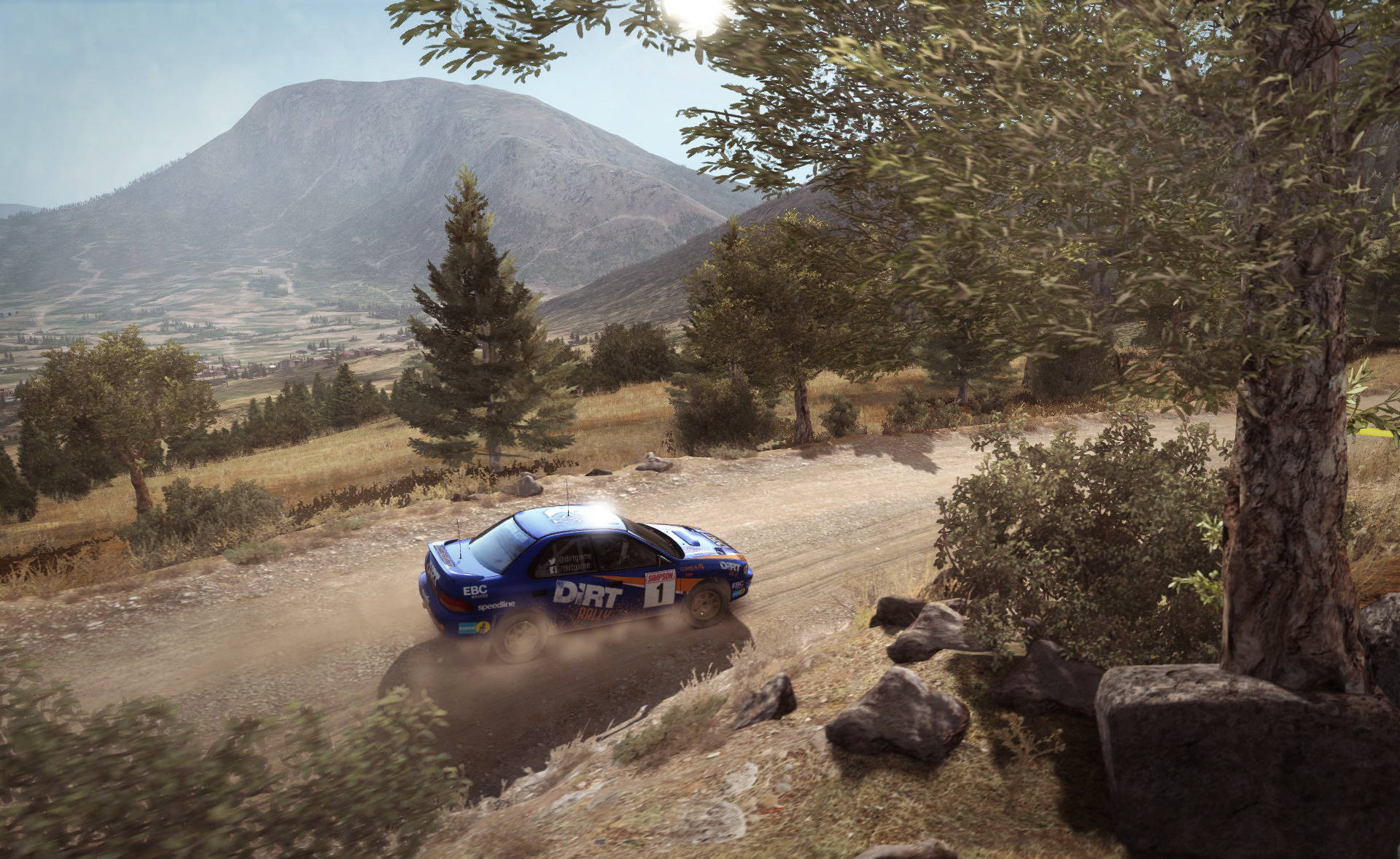 Dirt Rally 速霸陸行駛於鄉間路 壁紙 — 一輛藍色拉力賽車在陽光明媚的山區景觀中沿著蜿蜒的泥土路疾馳，揚起灰塵。