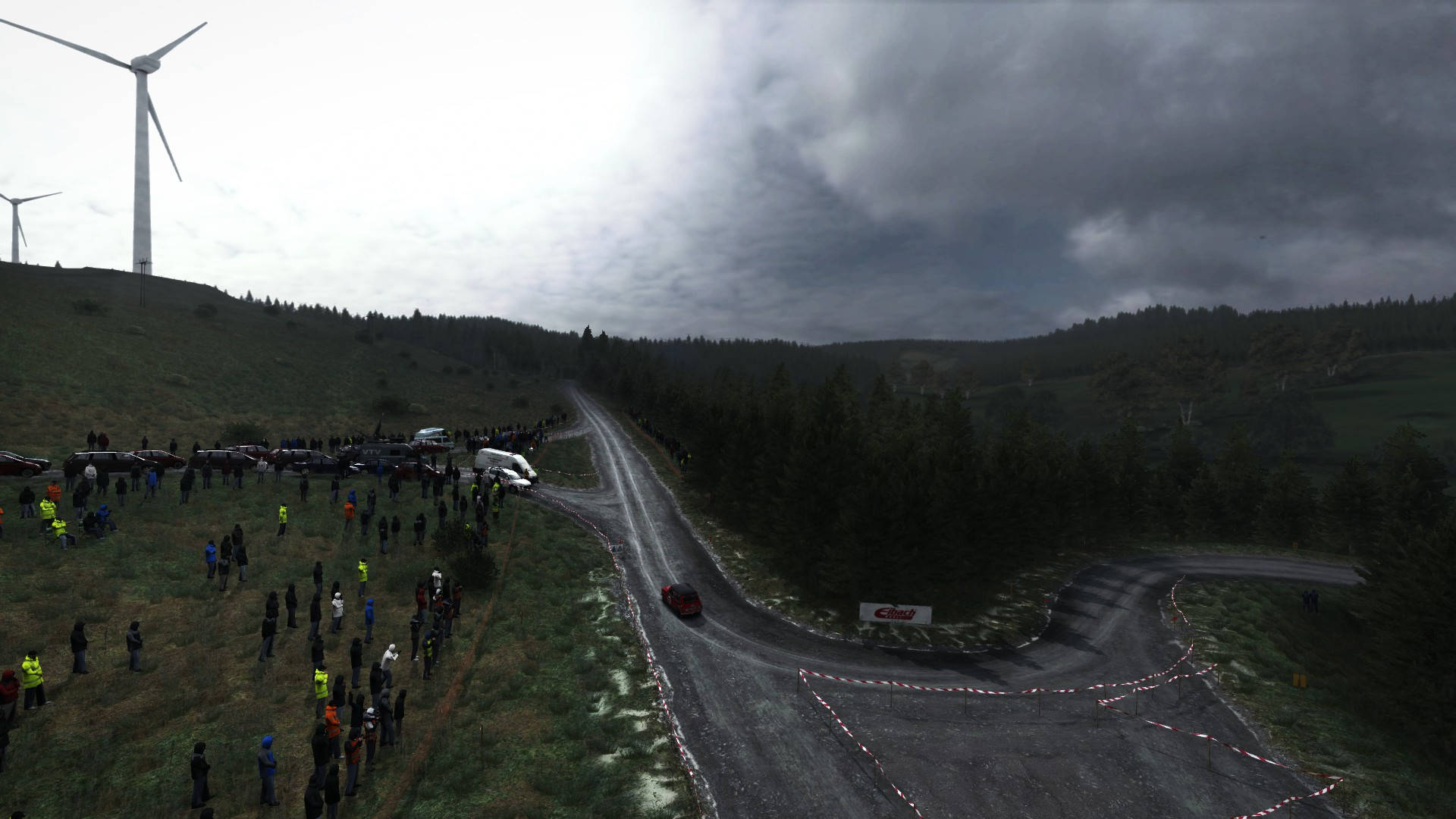 《Dirt Rally》賽道 壁紙 — 一輛紅色拉力賽車在泥濘的森林賽道上飛馳，觀眾沿路邊排列，陰天背景下遠處有風力發電機。