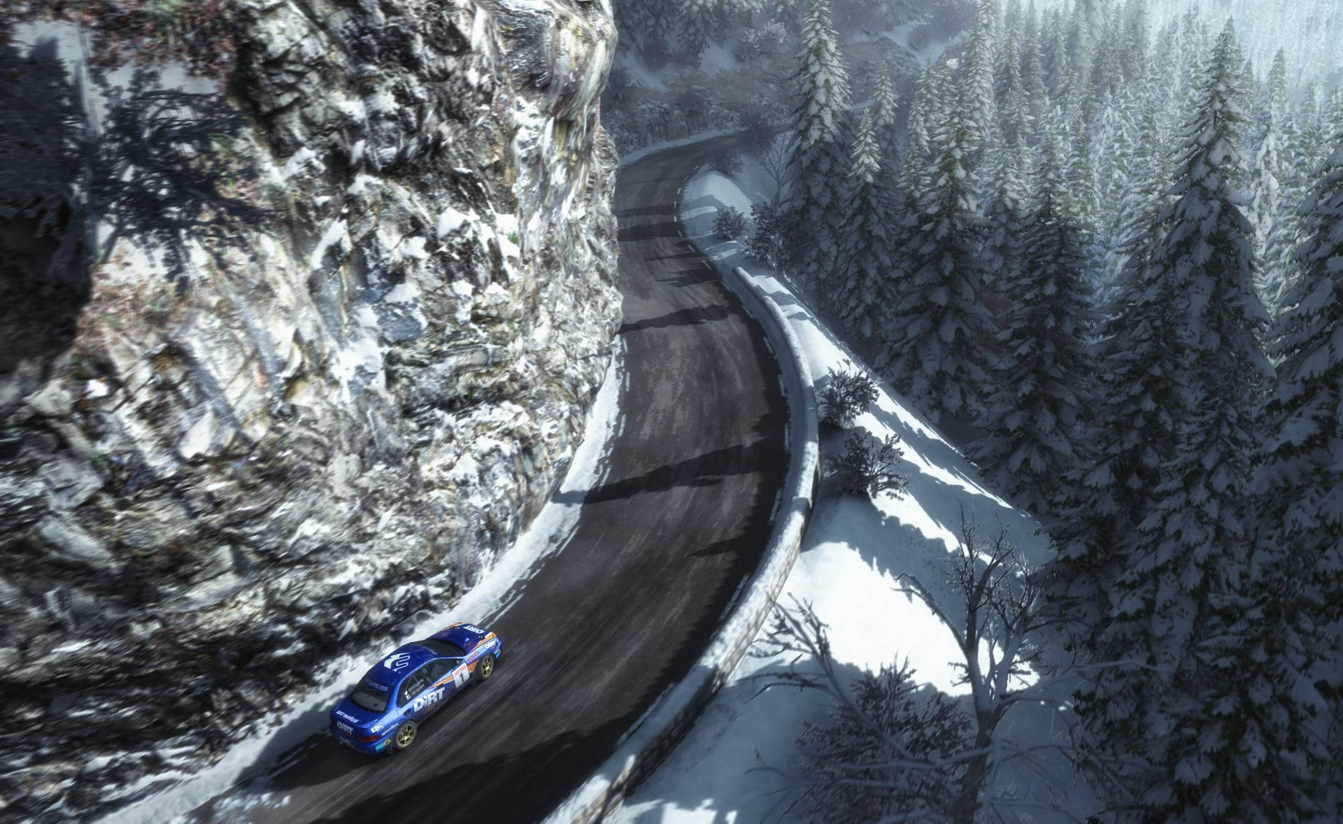 Dirt Rally 賽車行駛於山區 壁紙 — 一輛藍色拉力賽車沿著蜿蜒的雪山道路飛馳，兩旁是白雪覆蓋的松樹和陡峭的岩石懸崖。