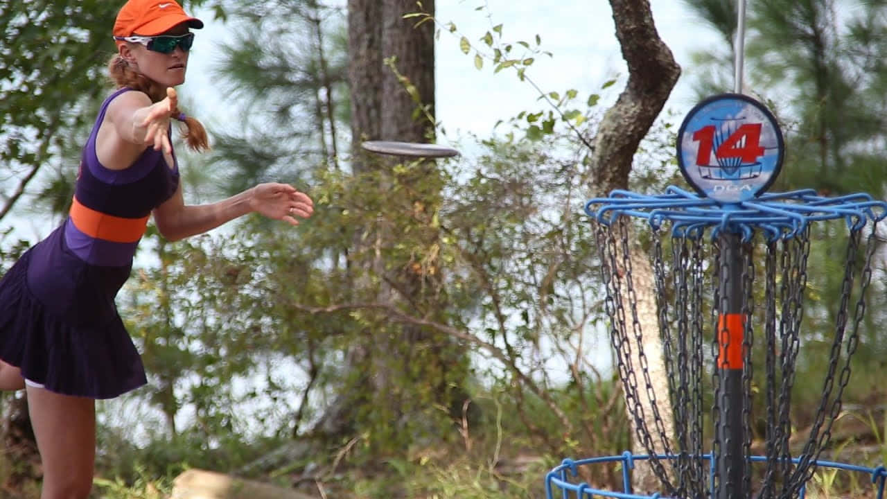 Disc Golf Hybrid 最佳 Ultimate Frisbee 背景 壁紙 — 一位穿著紫色連衣裙和橙色帽子的女性，在樹林環繞的戶外環境中，將飛盤擲向一個標有14號的藍色飛盤高爾夫籃框。