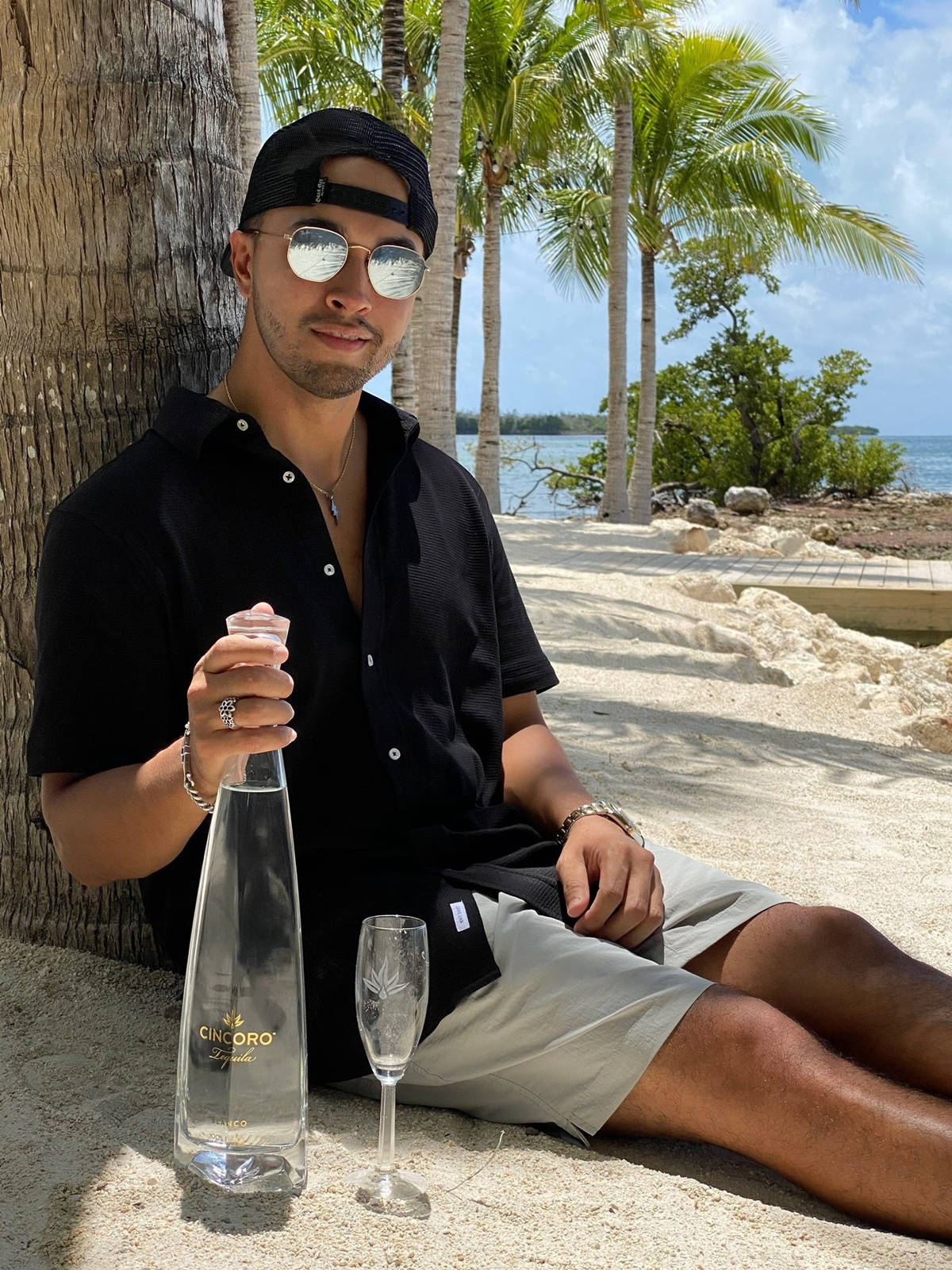 男子在棕櫚樹下拿著龍舌蘭酒瓶和酒杯在海灘上放鬆。
