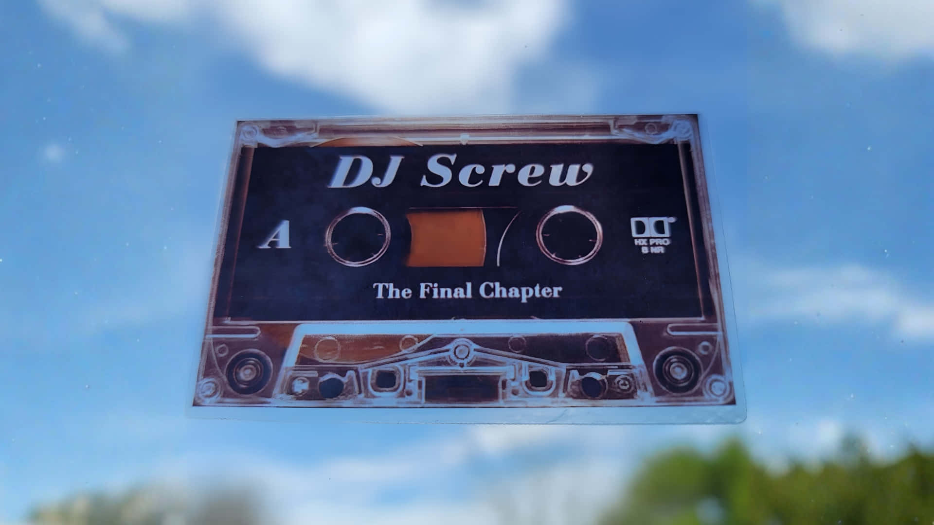 DJ Screw 最終章 壁紙 — 一張復古的DJ Screw卡式錄音帶貼紙，上面有'The Final Chapter'文字，在明亮的藍天和蓬鬆的雲朵中反射。