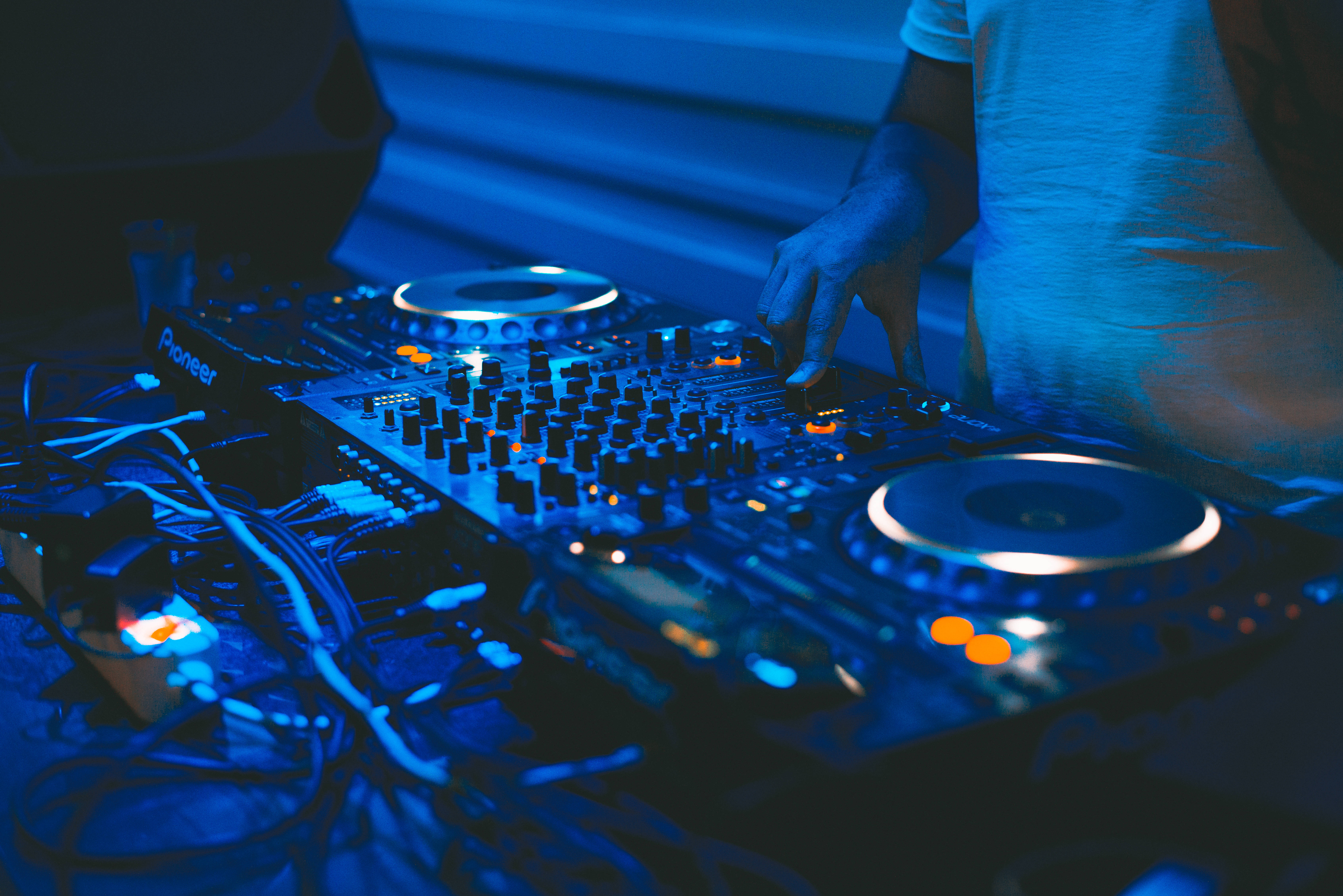 DJ 台前混音音樂 壁紙 — DJ的手正在調整Pioneer混音器上的旋鈕，混音器沐浴在冷藍色舞台燈光和發光的橙色點綴中。